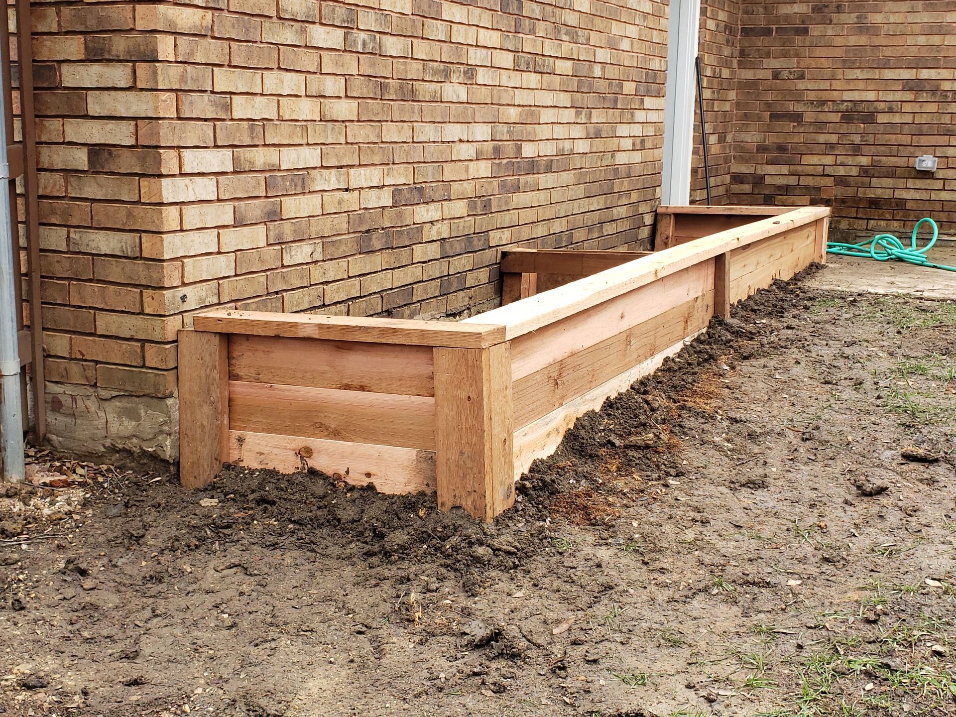 Wooden raised garden bed against a brick wall, filled with soil.