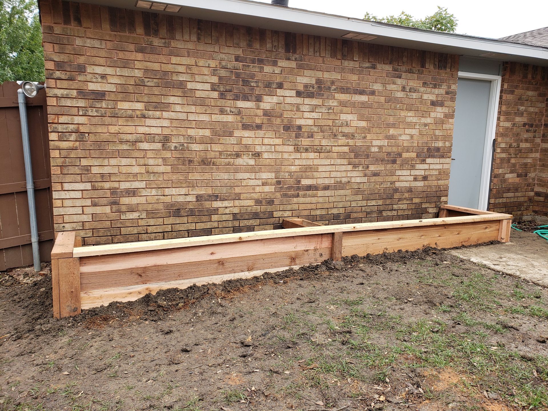 Wooden raised garden bed against a brick building; partially filled with soil.