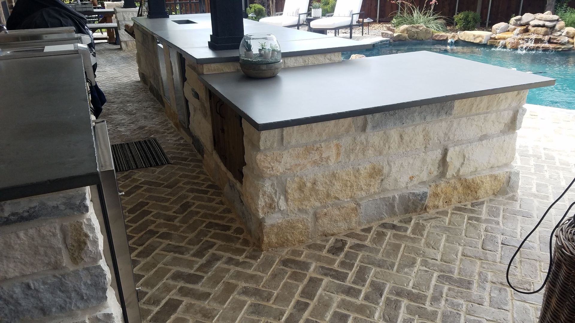 Outdoor kitchen with stone base and dark countertop next to a pool. Brick patio.