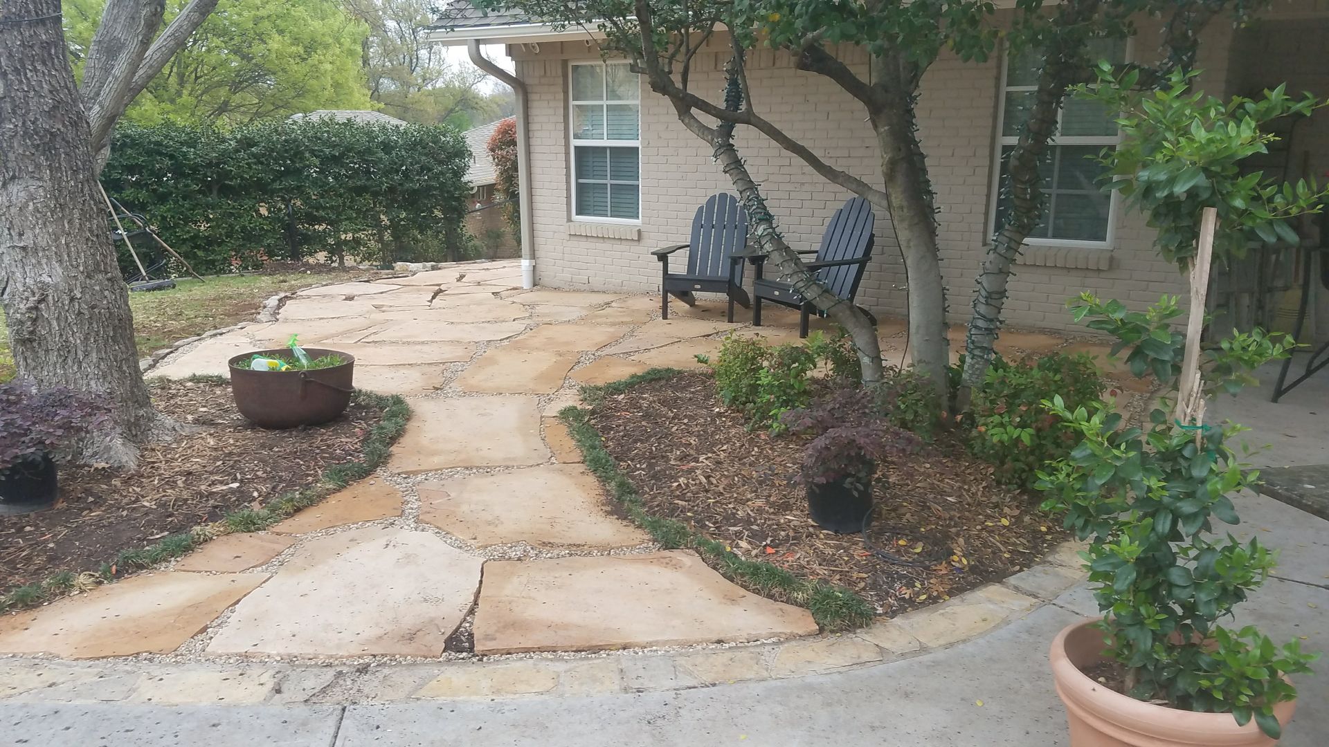 Flagstone patio with seating area beneath trees and a small brick building.