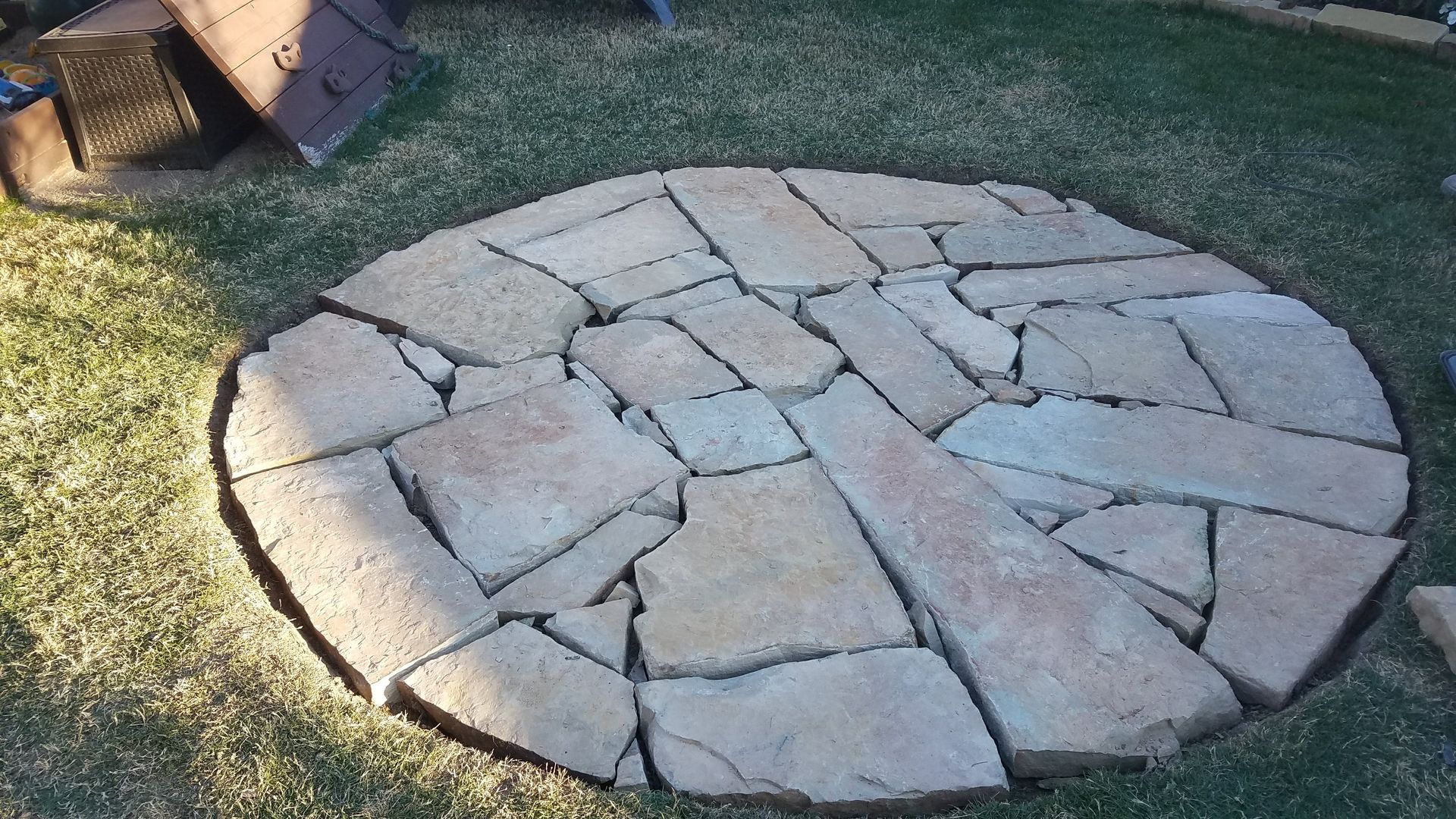 Circular patio made of irregularly shaped tan stones on green grass.