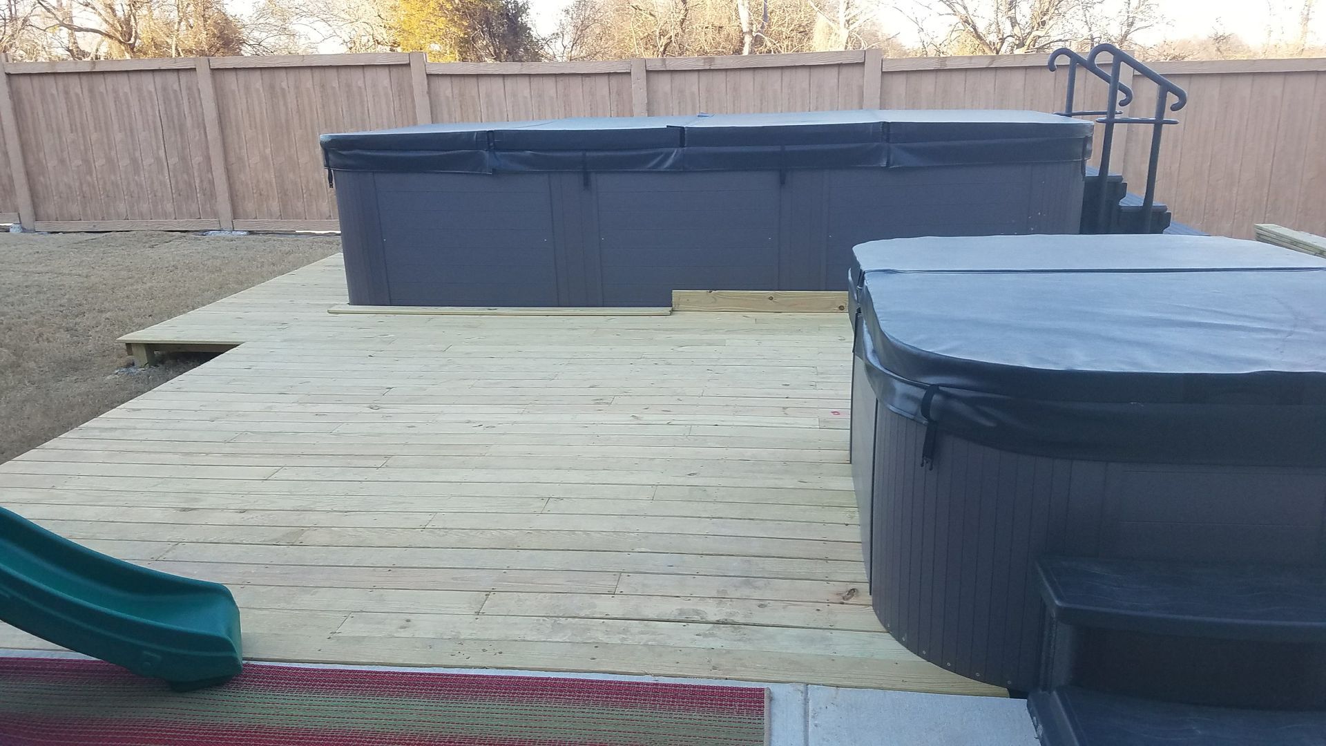 Deck with two hot tubs covered with black lids. A green slide is in view. Brown fence in background.