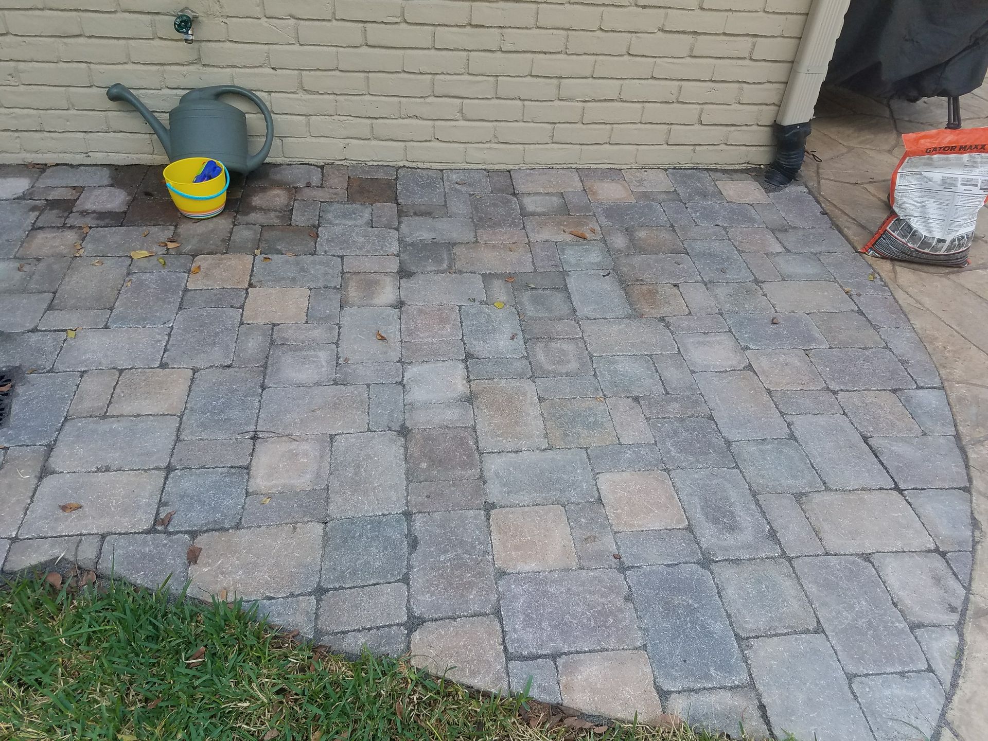 Brick patio with grass border, by a brick wall and a downspout; watering can and bucket are present.