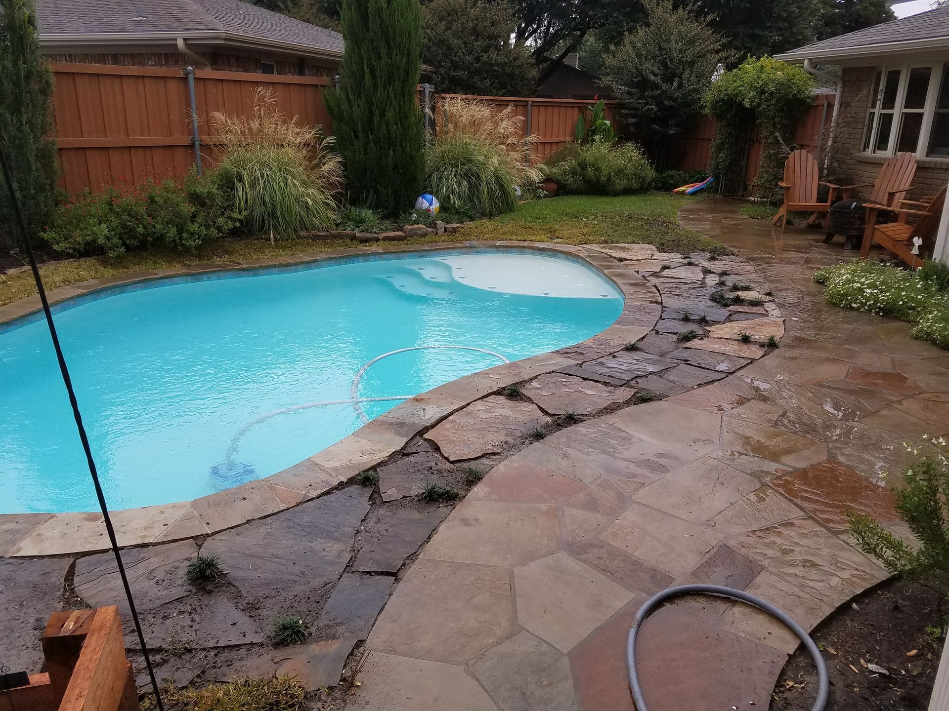 Swimming pool with stone patio in backyard; brown fence, green lawn and foliage.