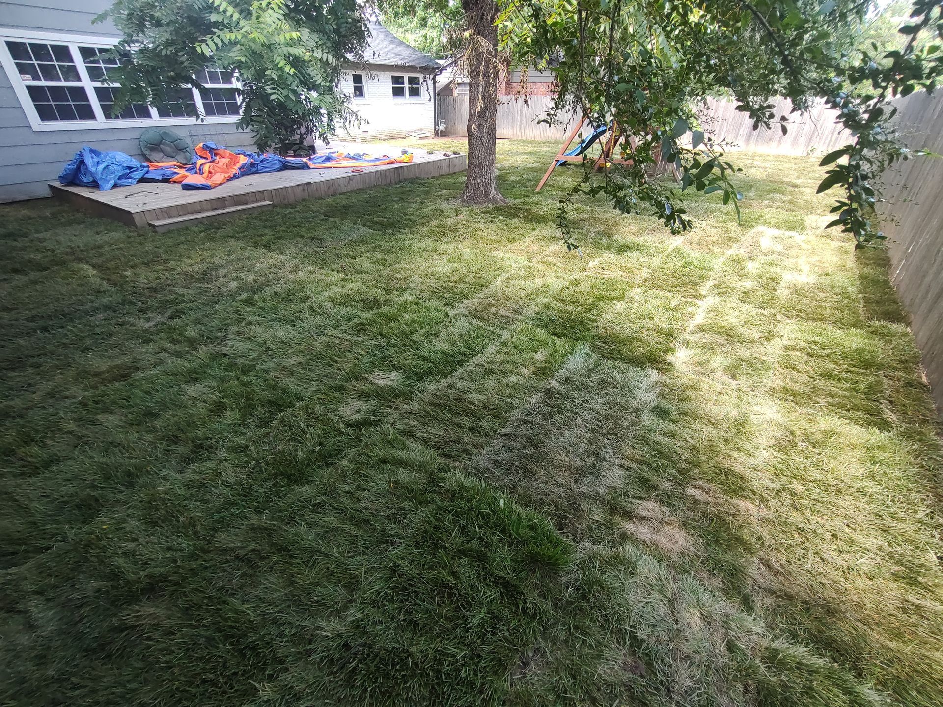 Backyard with patchy green grass, a patio, a fence, and a tree, under a bright sky.
