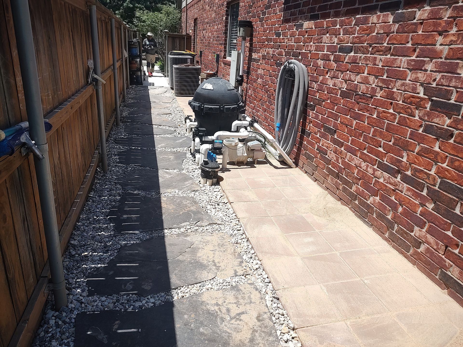 Narrow alleyway between a wooden fence and brick wall. Pool equipment sits on pavers.