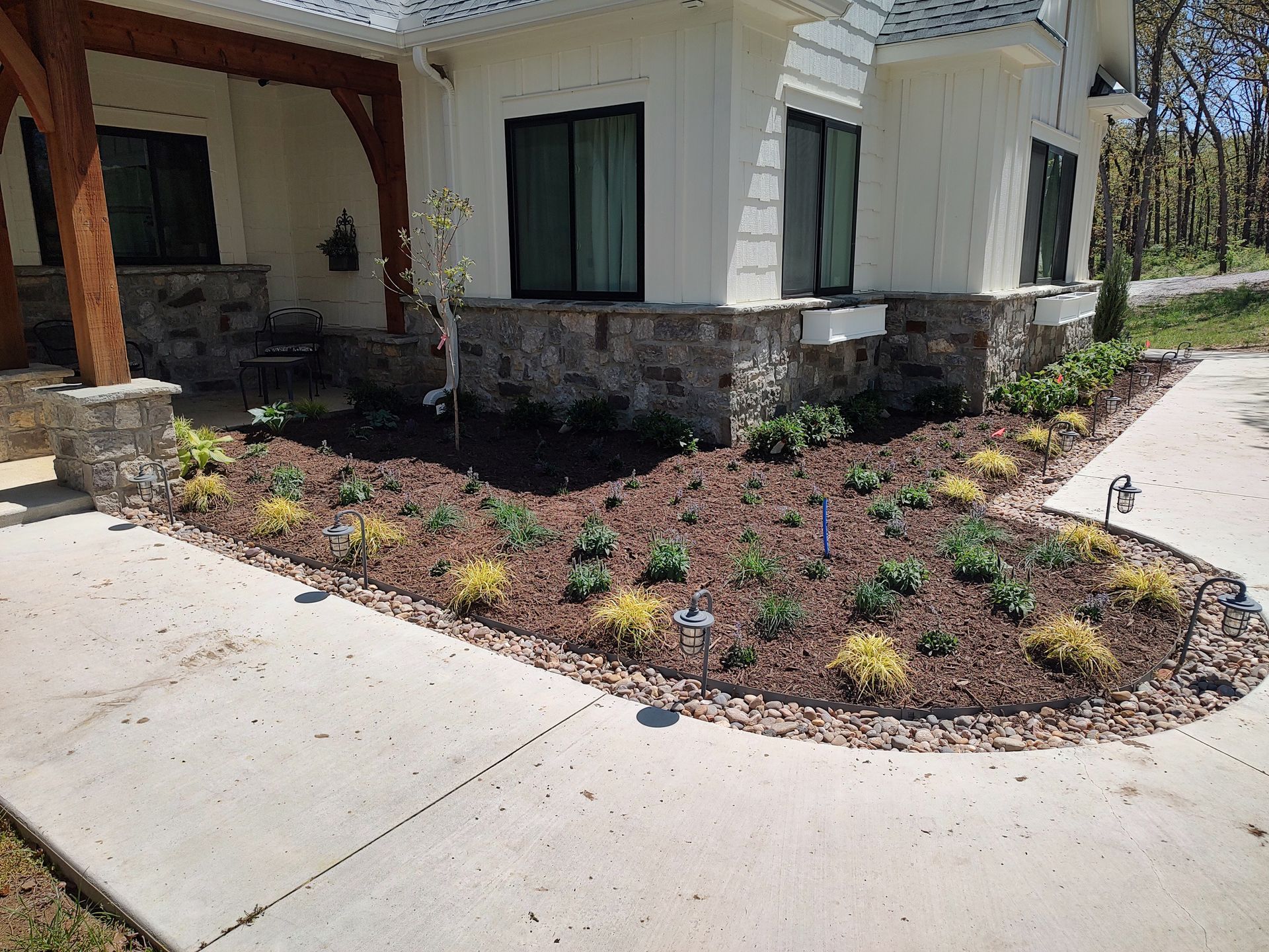 Landscaped flower bed with various plants and pathway lights in front of a white house with stone accents.