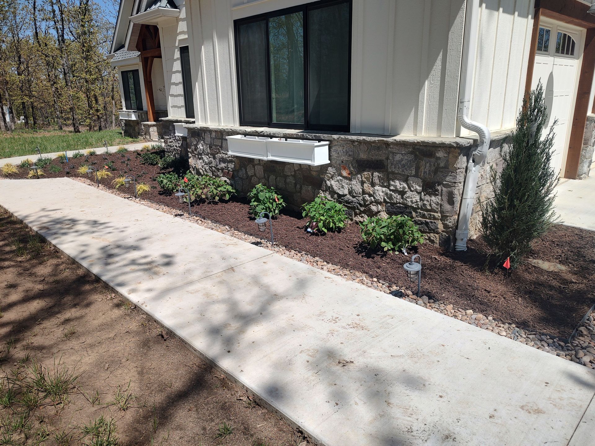 Sidewalk leads to a house with a stone foundation, window box, and landscaped flower bed.
