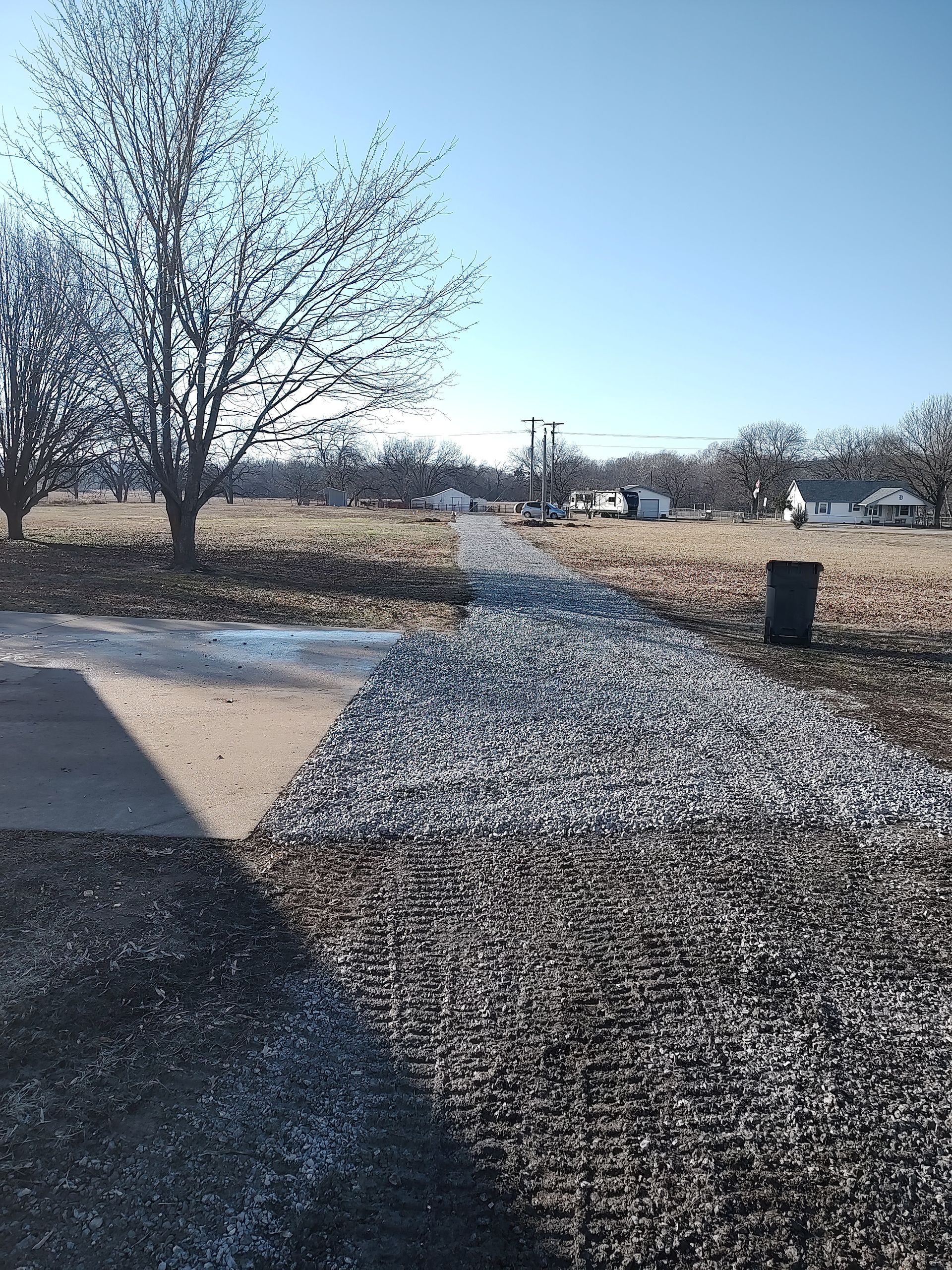 Gravel path leads through a field with scattered trees under a clear, sunny sky. A trash can is to the right.