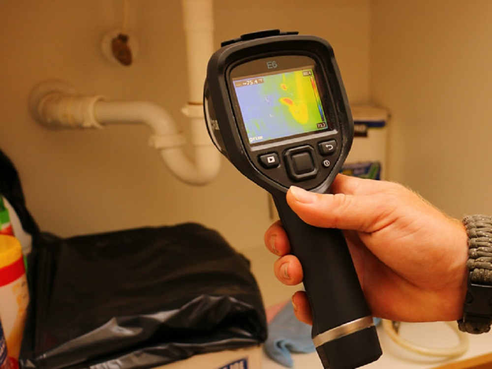 A person uses a thermal imaging camera under a sink to detect water leaks in plumbing.