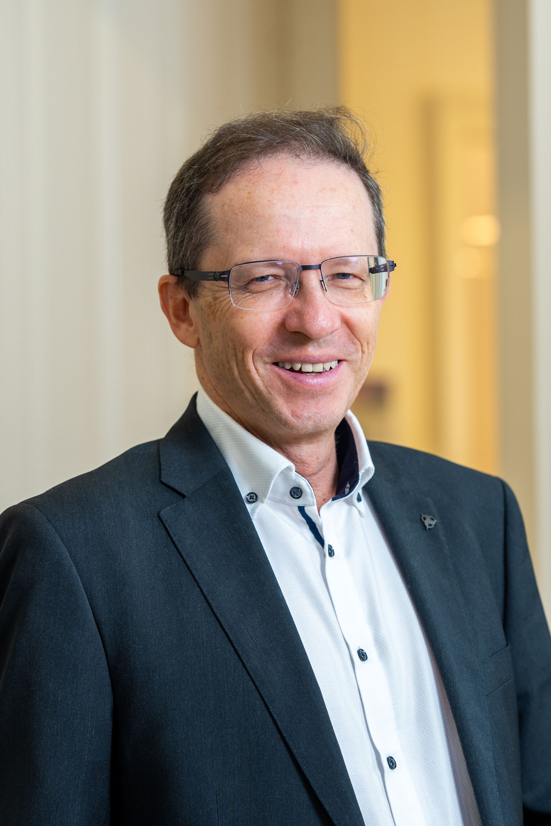 Mann mit Brille, schwarzem Anzug und roter Krawatte lächelt in einem Büro stehend.