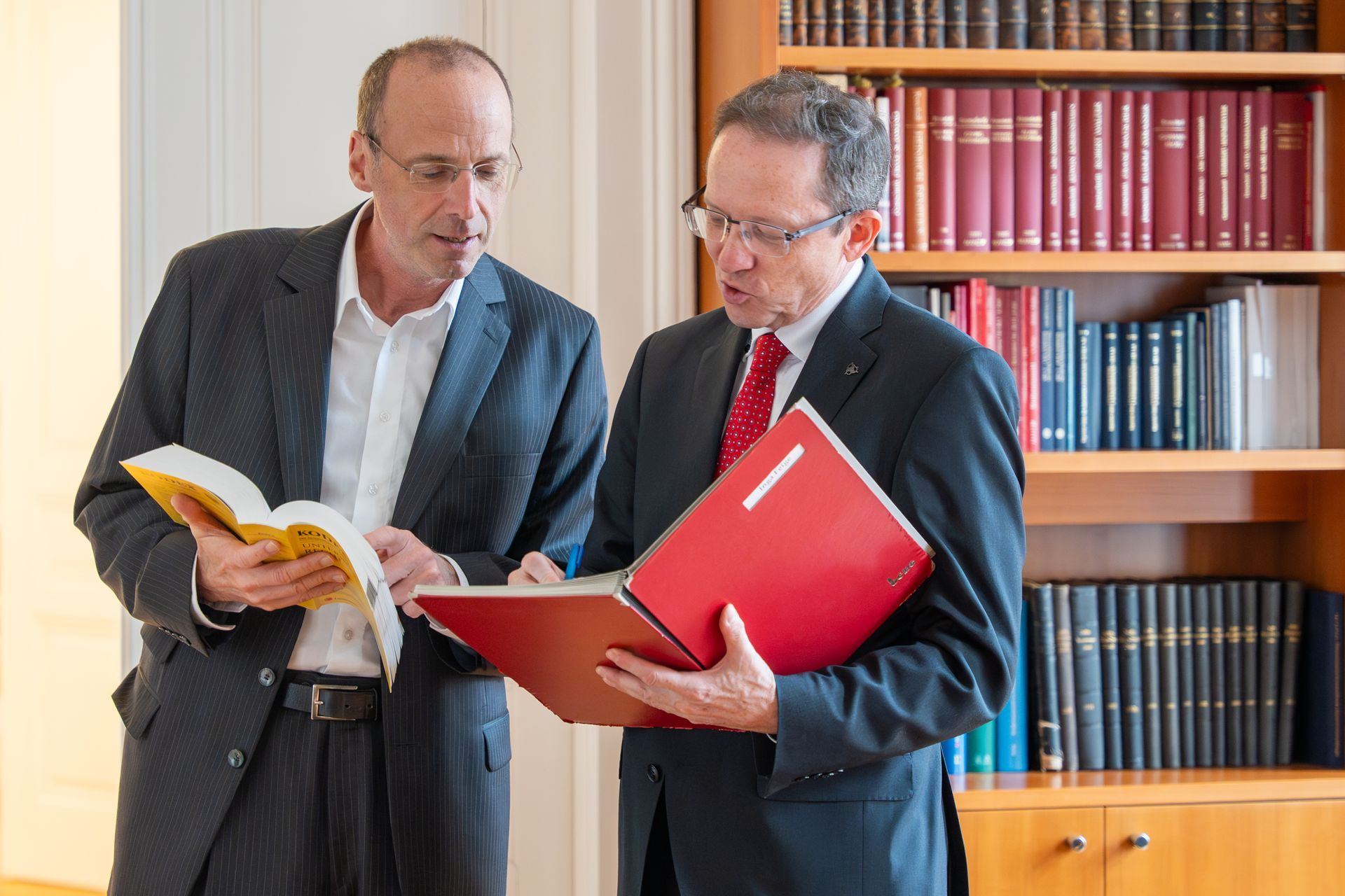Zwei Männer in Anzügen sichten Dokumente in der Nähe eines Bücherregals; einer hält ein Buch in der Hand.