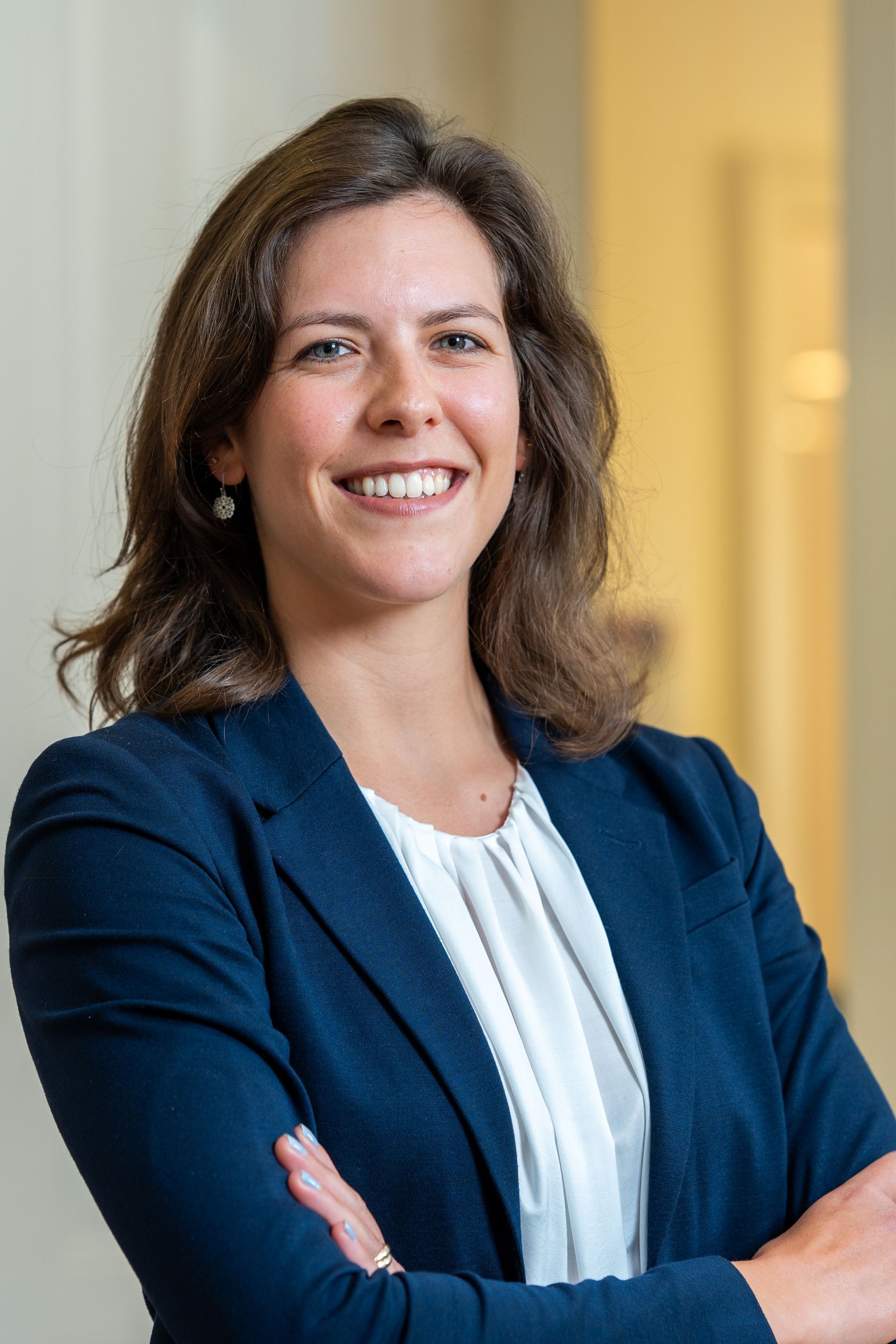 Frau in marineblauem Blazer und weißer Bluse, lächelnd mit verschränkten Armen, in einem hell erleuchteten Büro.