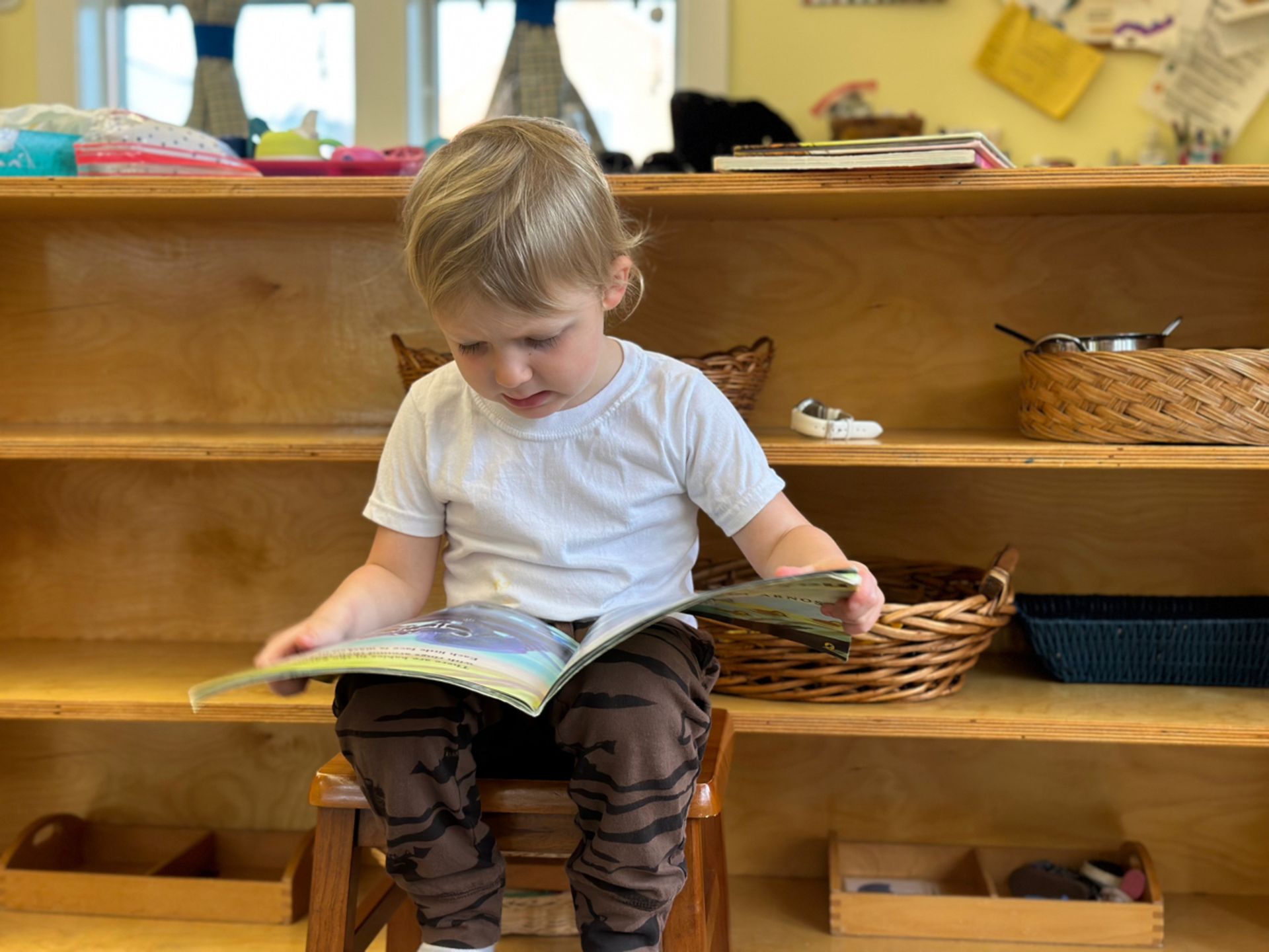 A Montessori  young boy sits on a stool reading a book