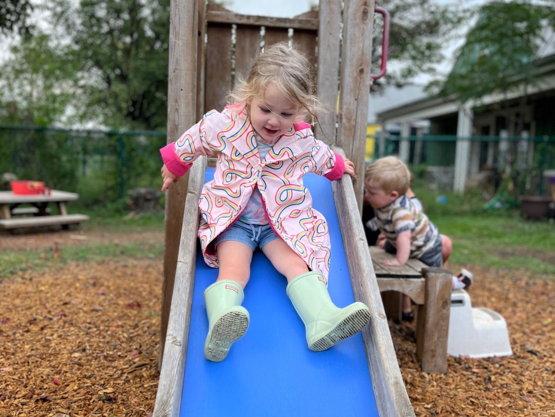 A Montessori little girl wearing rain boots is sitting on a slide