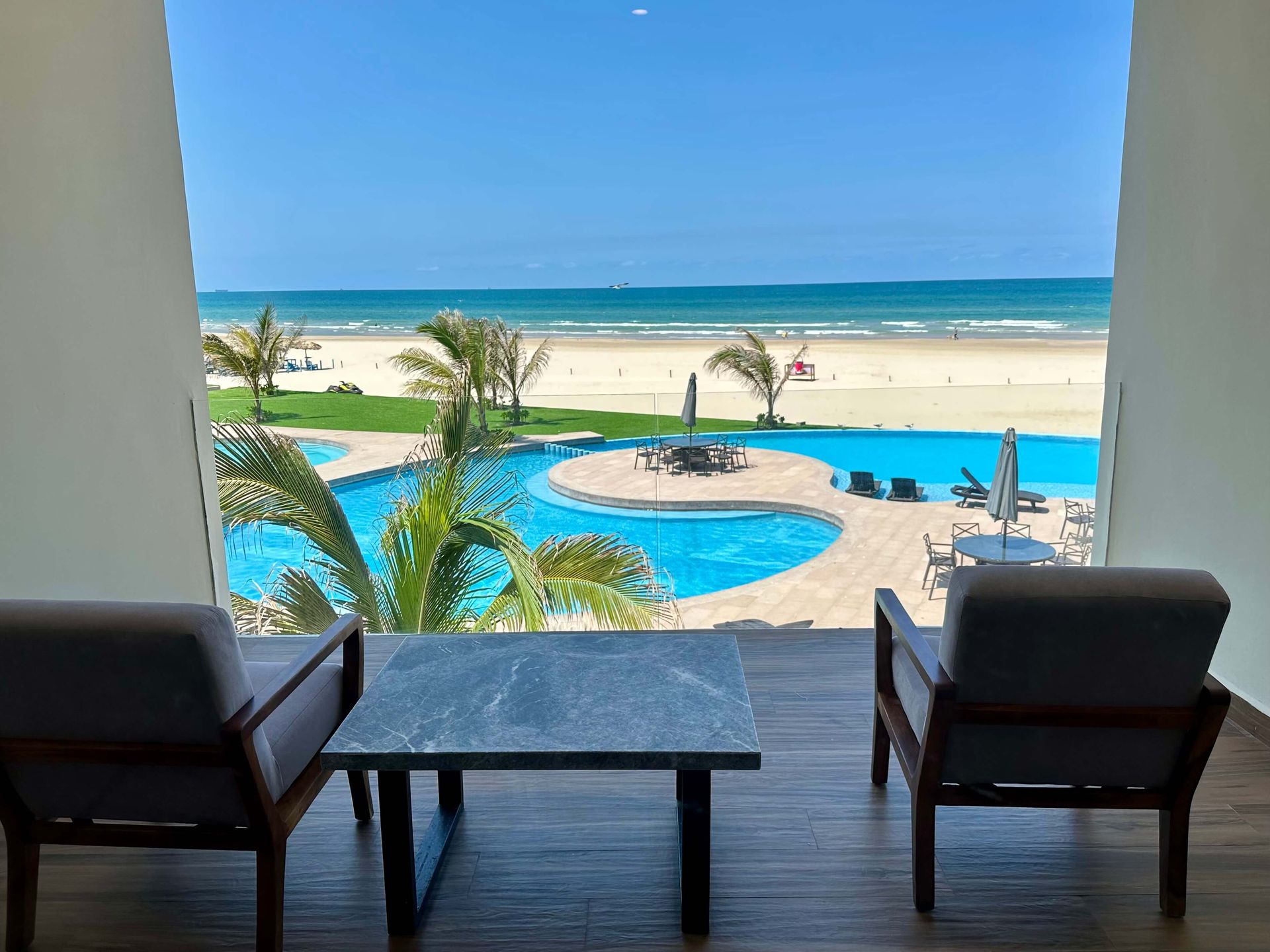 A room with two chairs and a table with a view of a swimming pool and beach.