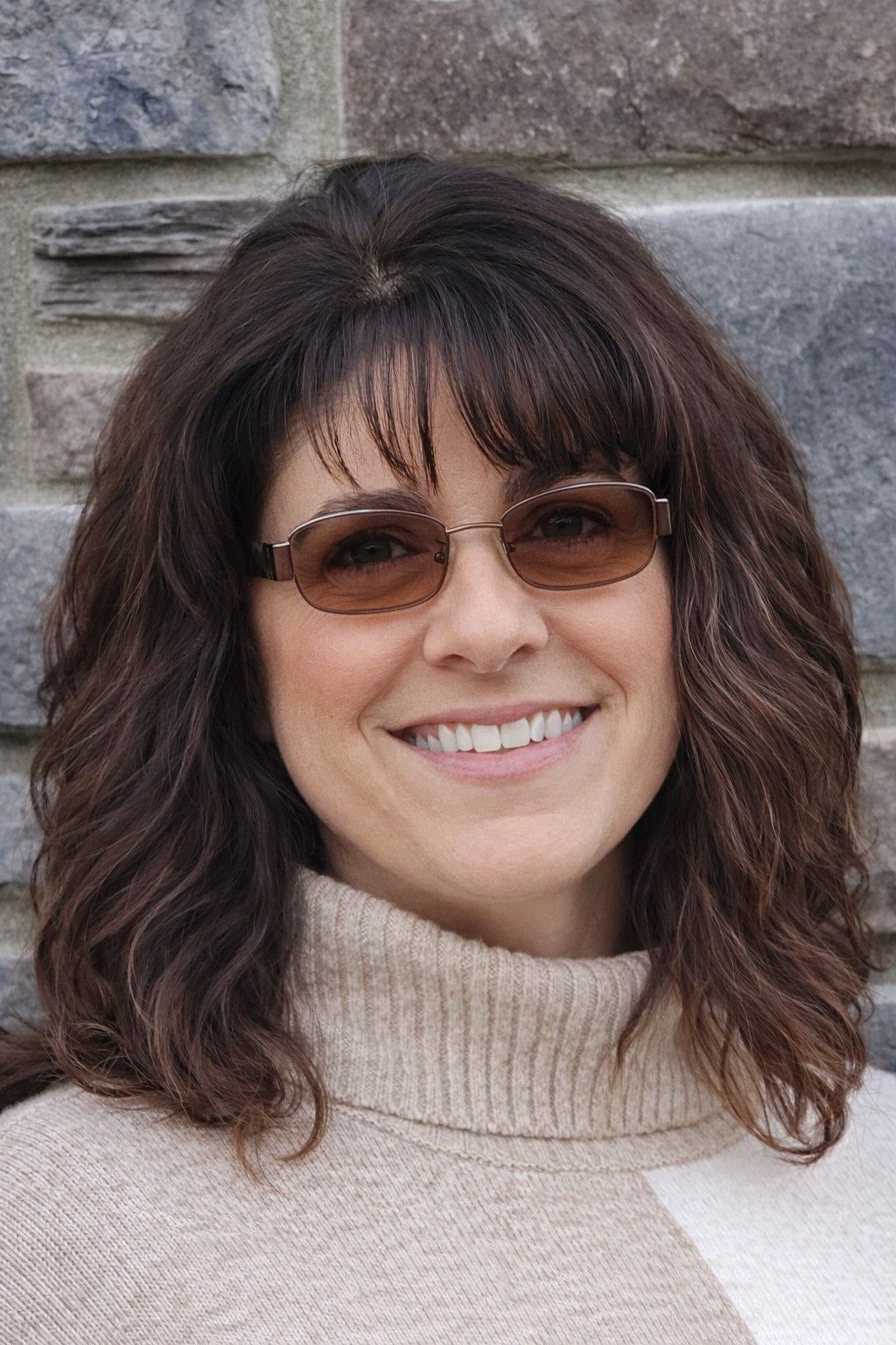 Woman wearing glasses smiles at the camera in front of a stone wall.