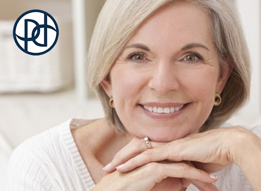 Smiling woman with short blonde hair, resting chin on hands. White top, small gold earrings.