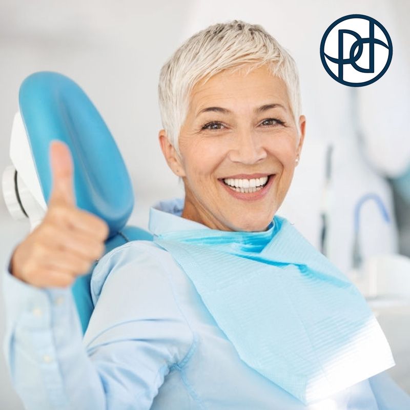 Woman in dentist chair smiling, giving thumbs up. Blue bib and chair.  White teeth and short, gray hair.