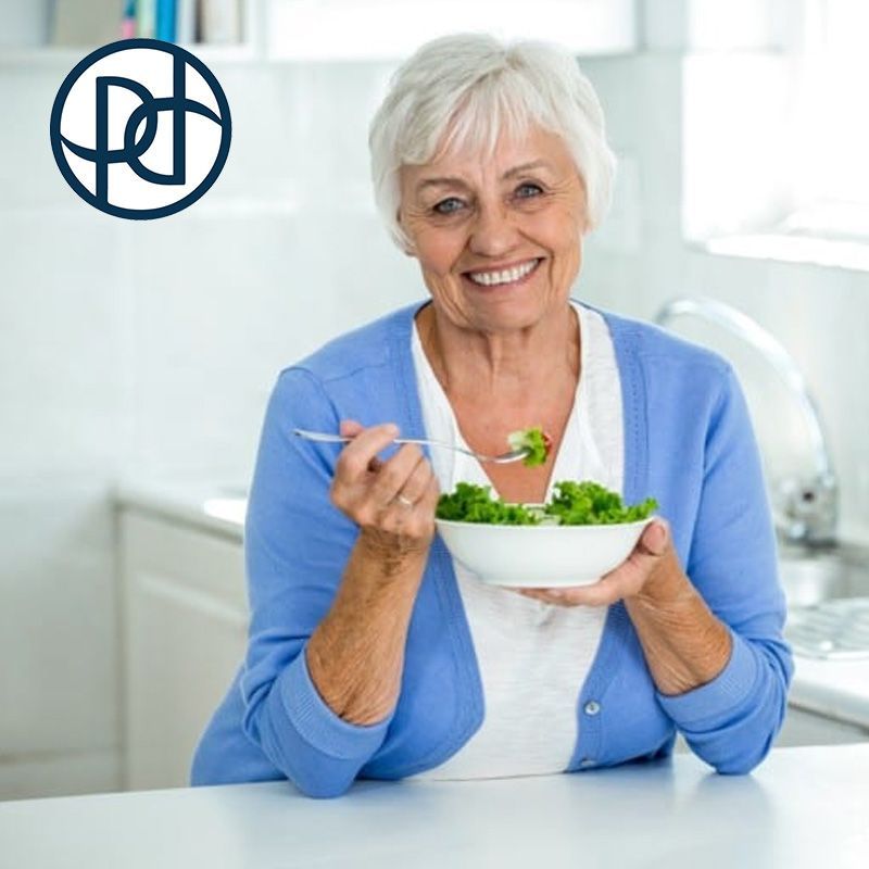 Smiling woman eating salad in a kitchen, wearing a blue cardigan.