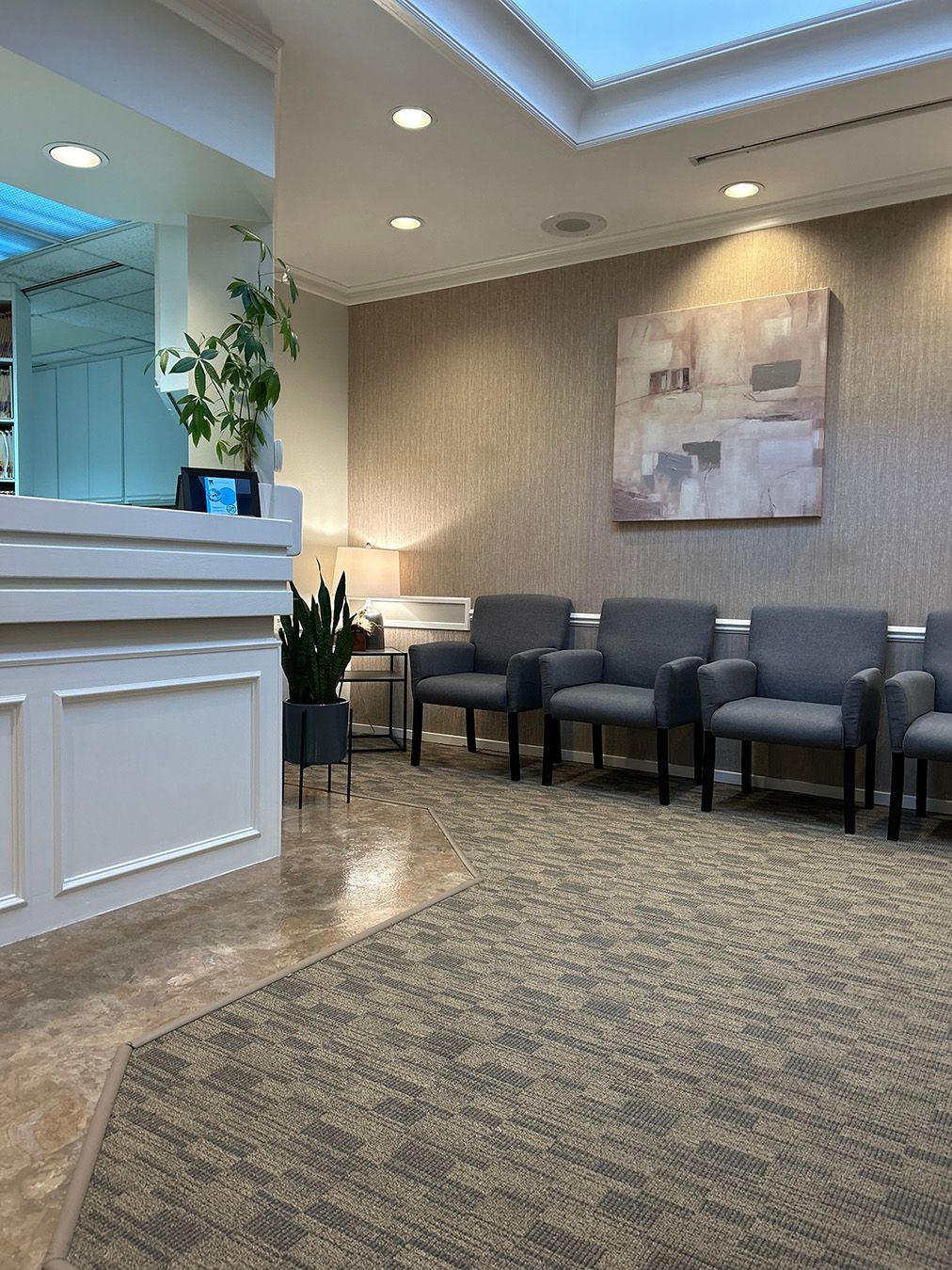 Reception area with chairs, desk, plants, and artwork. Neutral colors, natural light, and patterned carpet.