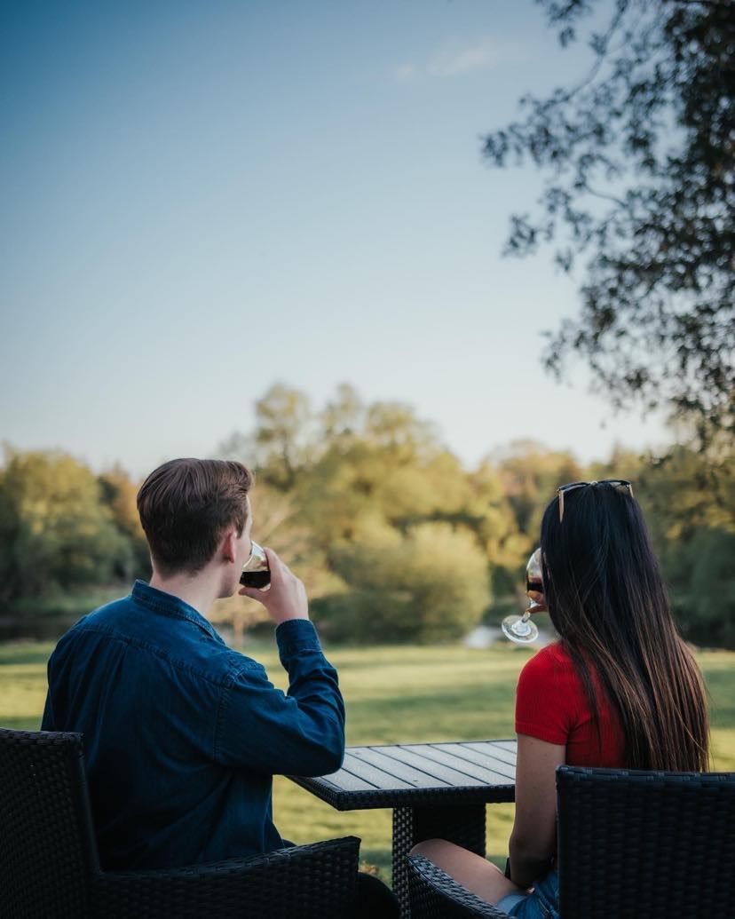 Couple drinking wine at an outdoor table, overlooking a green field and trees, under a blue sky.