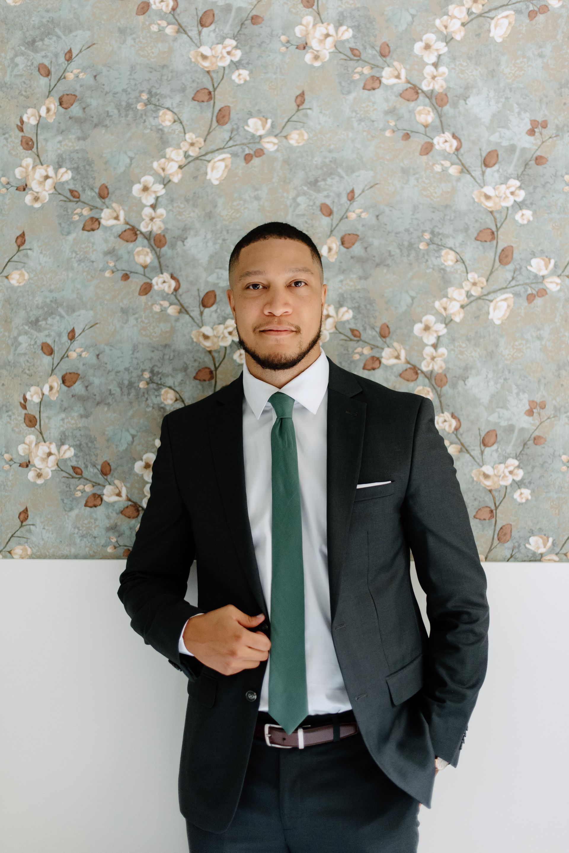 Man in a dark suit and green tie smiles against a floral patterned wall.