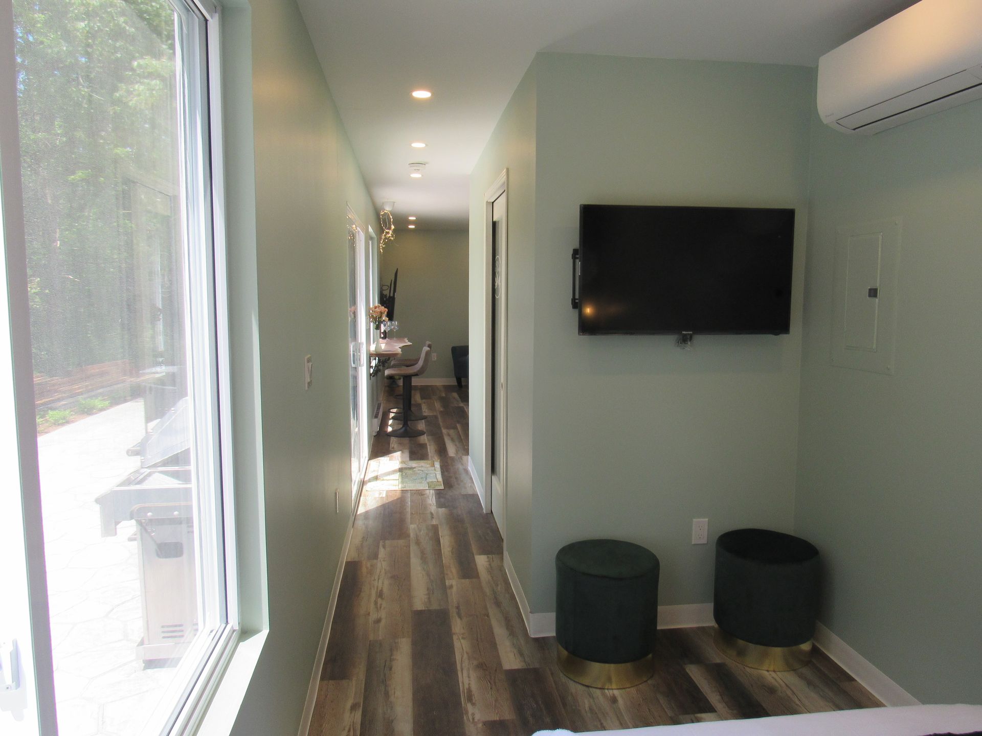Narrow room with wood floor, TV, two green stools, air conditioner, and large window.