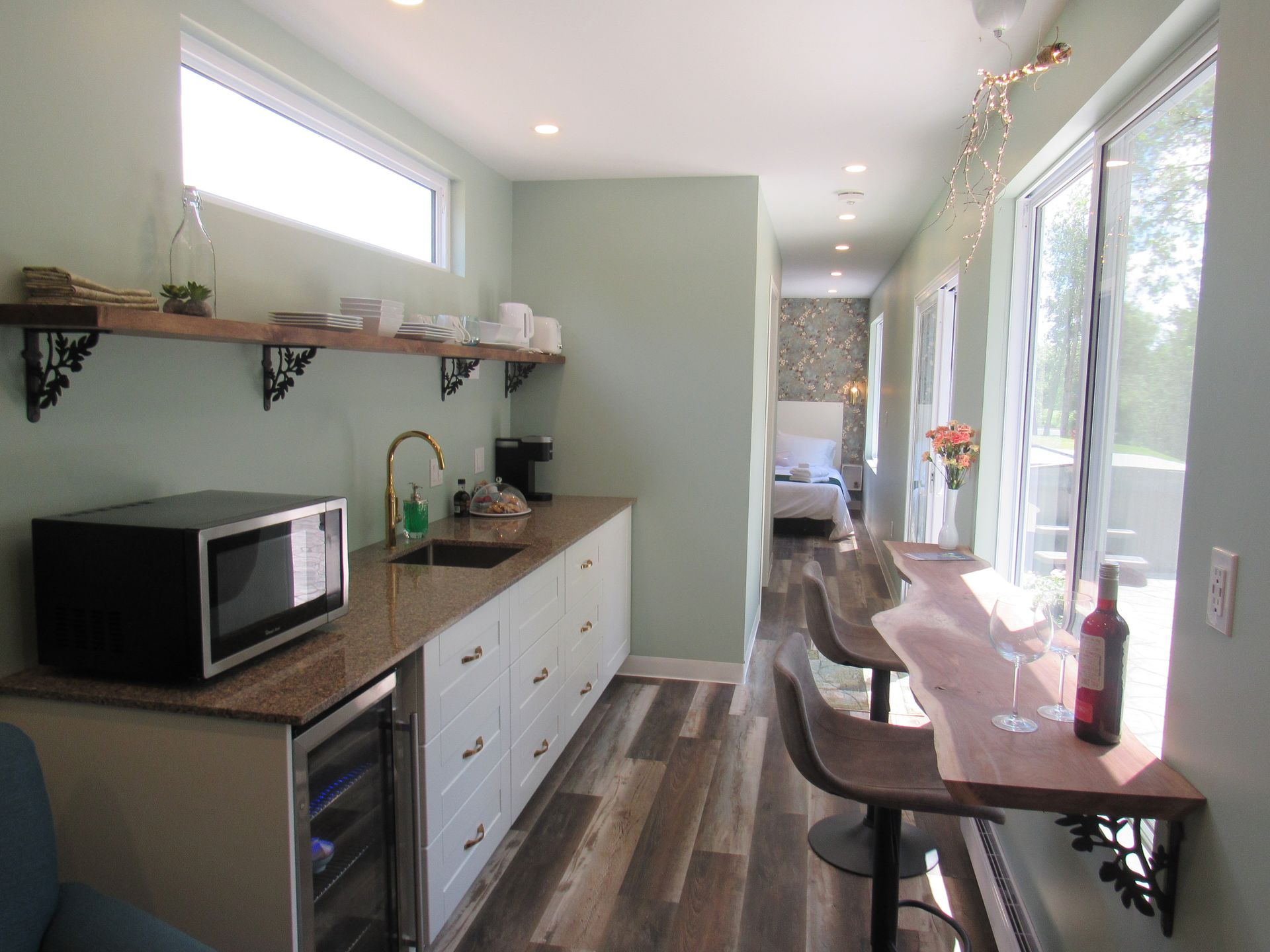 Interior view of a kitchenette with a microwave, sink, and wine bar, leading to a bedroom.