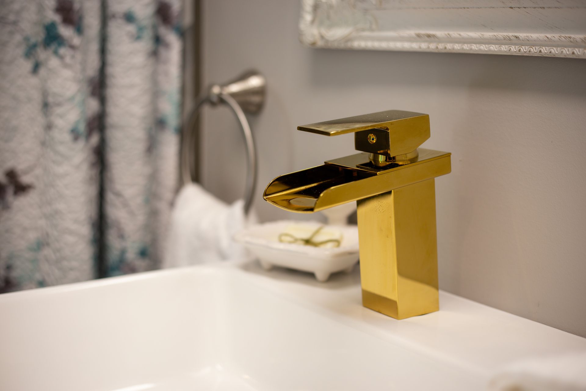 Gold faucet over a white sink in a bathroom, towel rack and floral curtain visible.