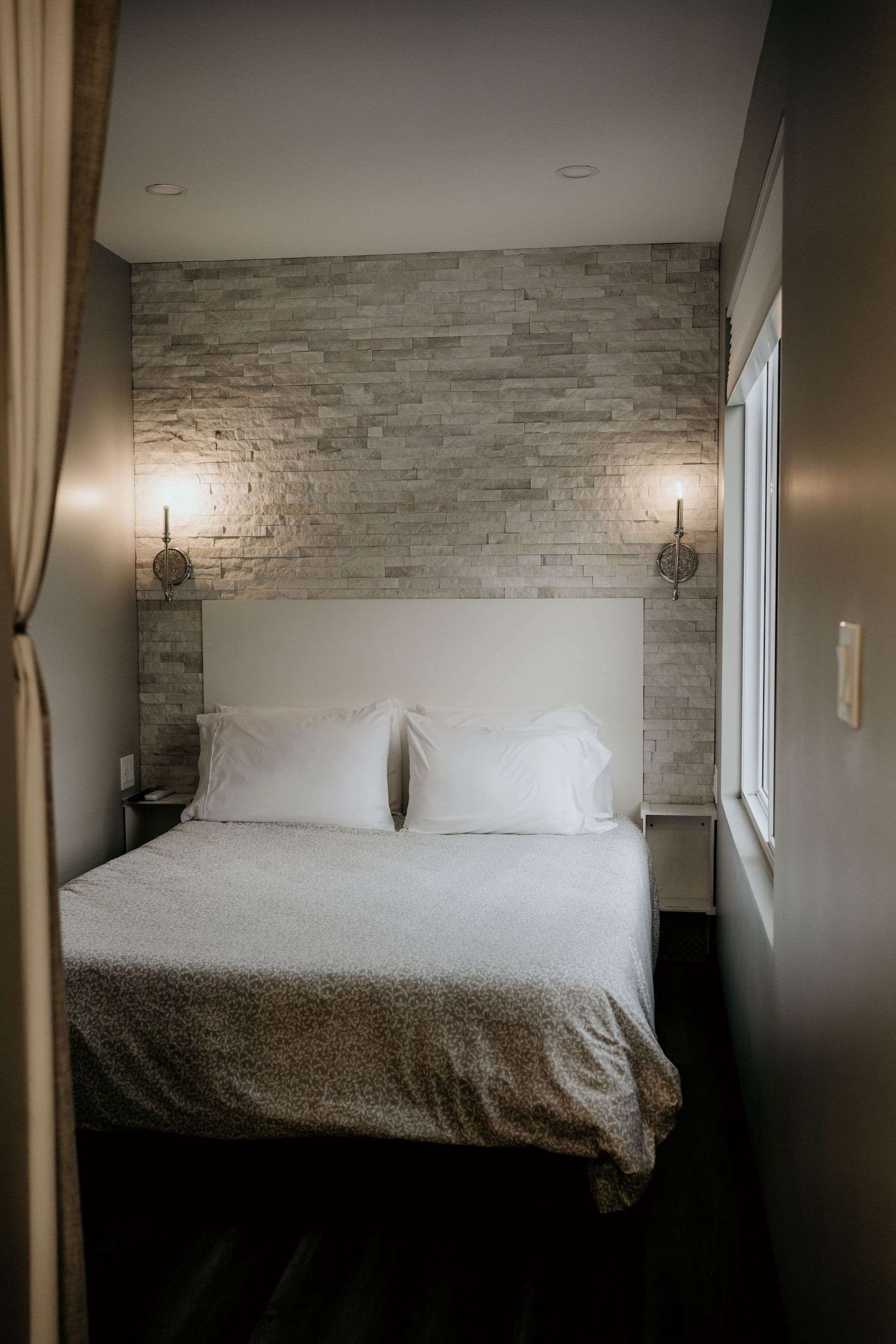 Bedroom with white bedding, brick accent wall, two sconces, and a window.