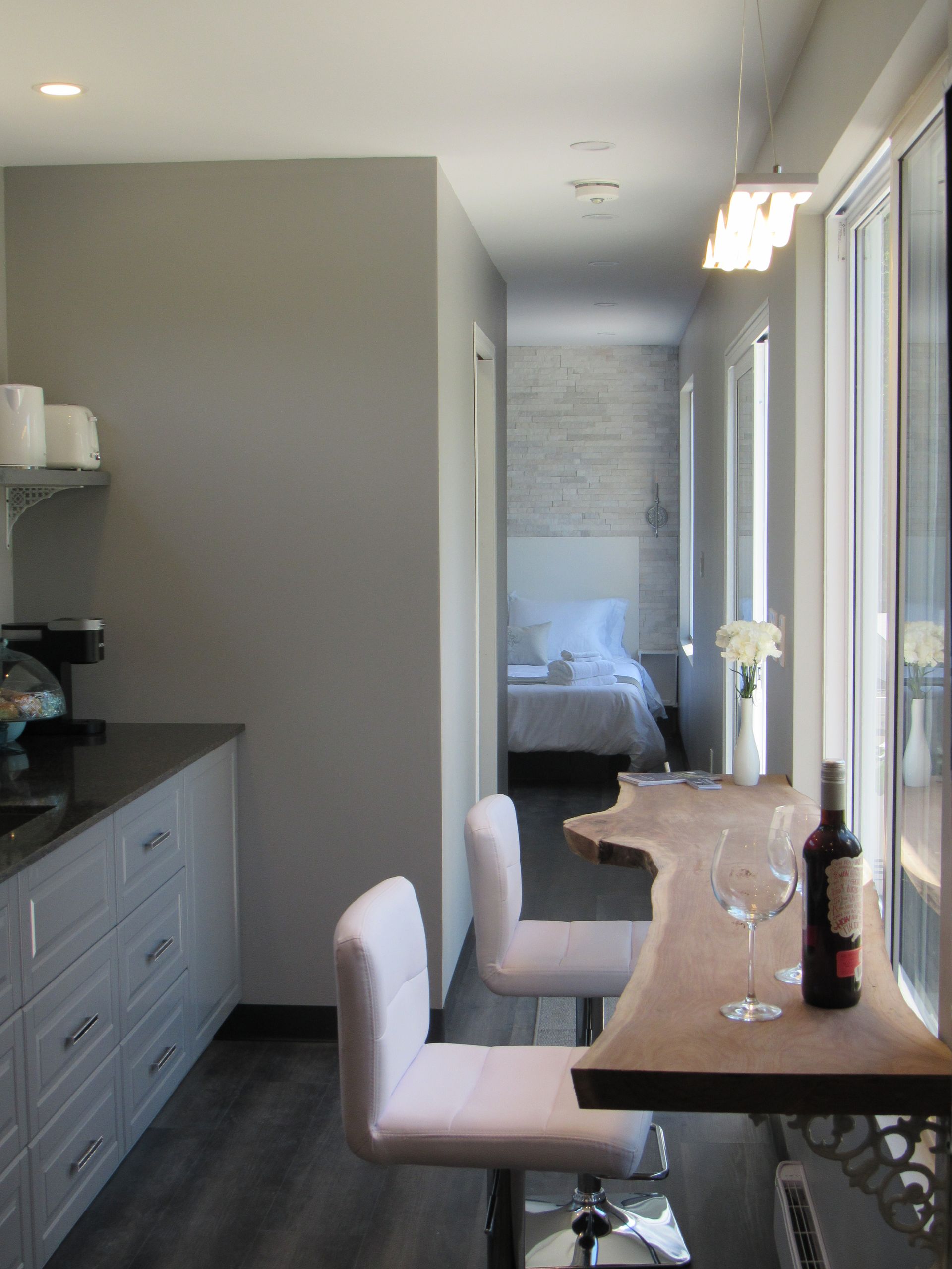 A modern, narrow room with a kitchen, bar, and bedroom visible. Gray walls, wood counter, pink bar stools.
