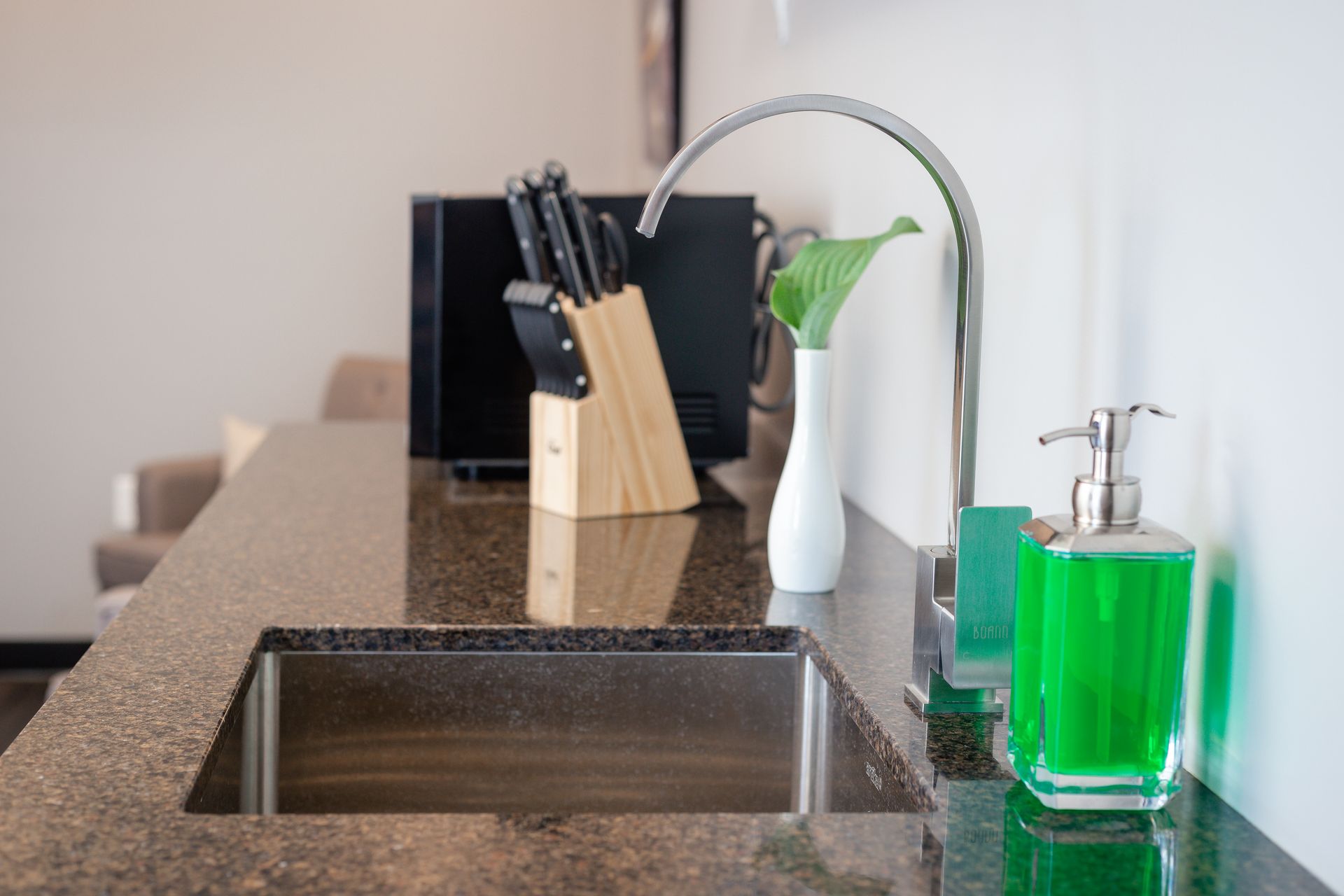 Kitchen sink with a faucet, knife block, soap dispenser, and a white vase with greenery on a dark countertop.