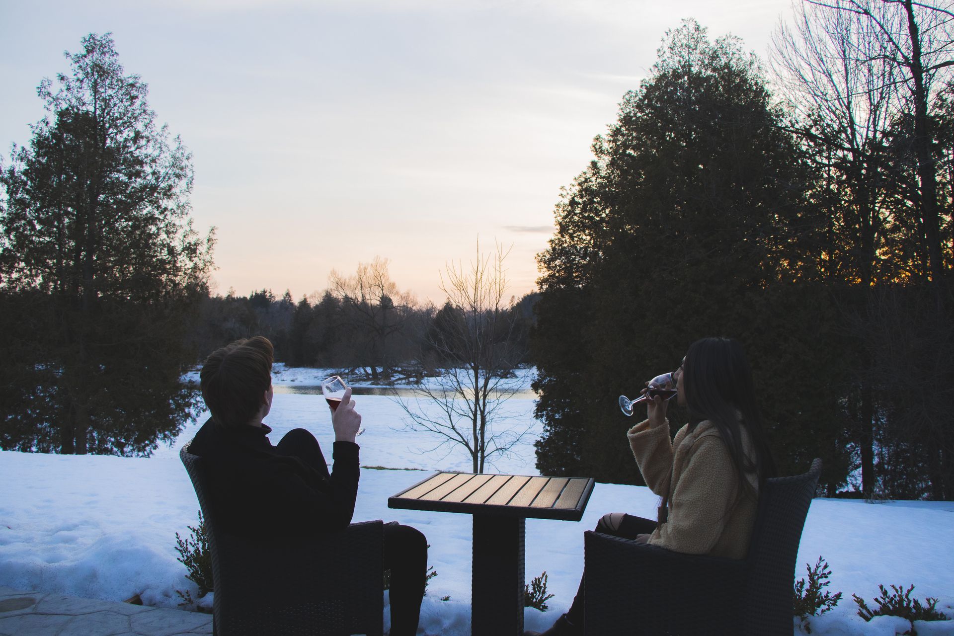 Couple toasting wine, seated at a table on a snowy patio, overlooking a serene, wintry landscape at sunset.