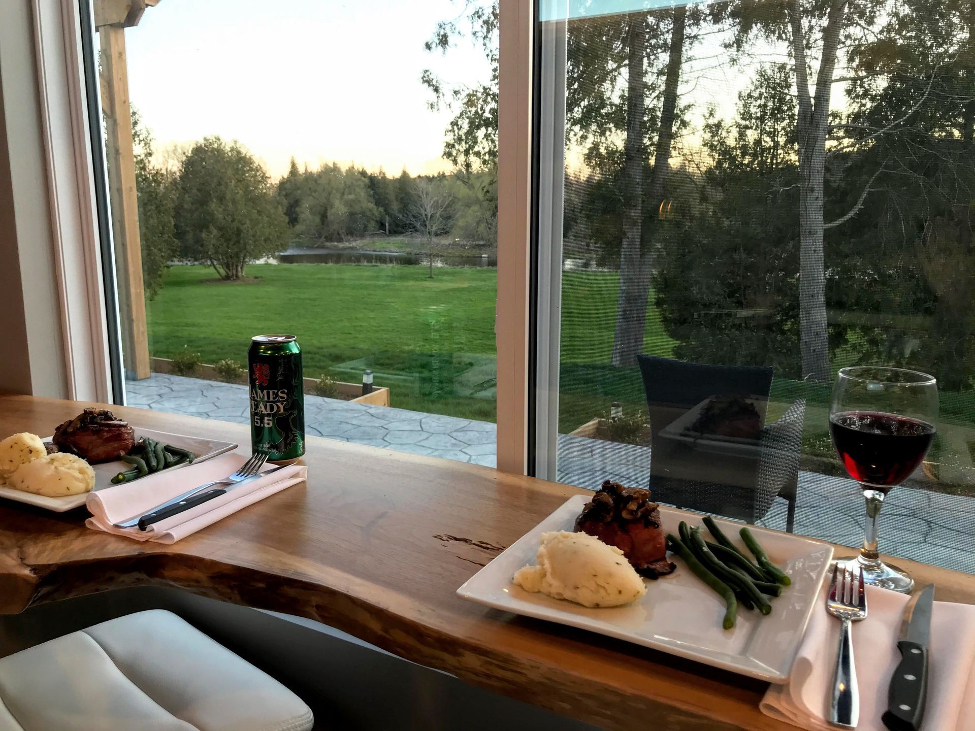Meal set on a wood counter with scenic backyard view; steak, mashed potatoes, green beans, beer, and wine.