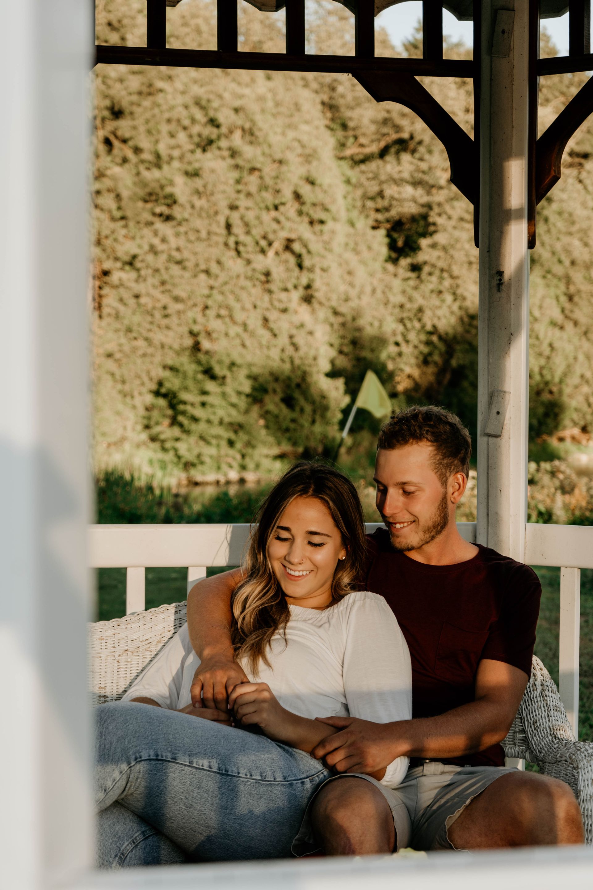 Couple in a gazebo, hugging and smiling. She's wearing a white shirt, he's wearing a maroon one. Outdoors, sunny day.