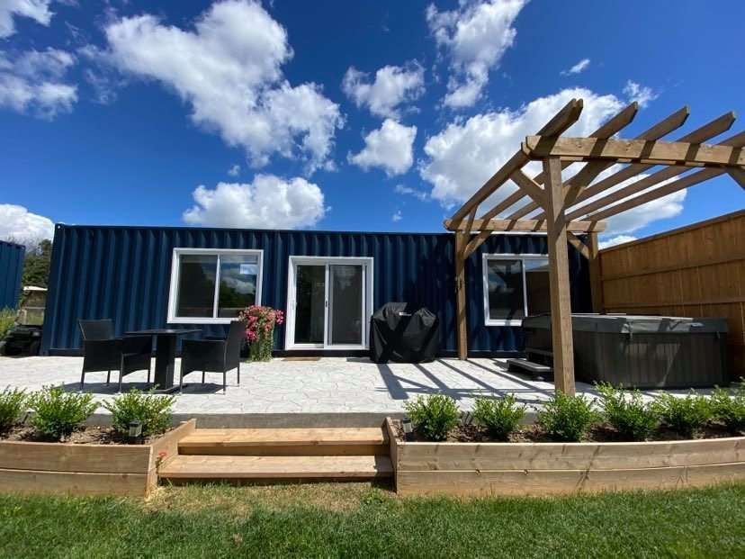 Blue shipping container home with patio, pergola, and hot tub on a sunny day.