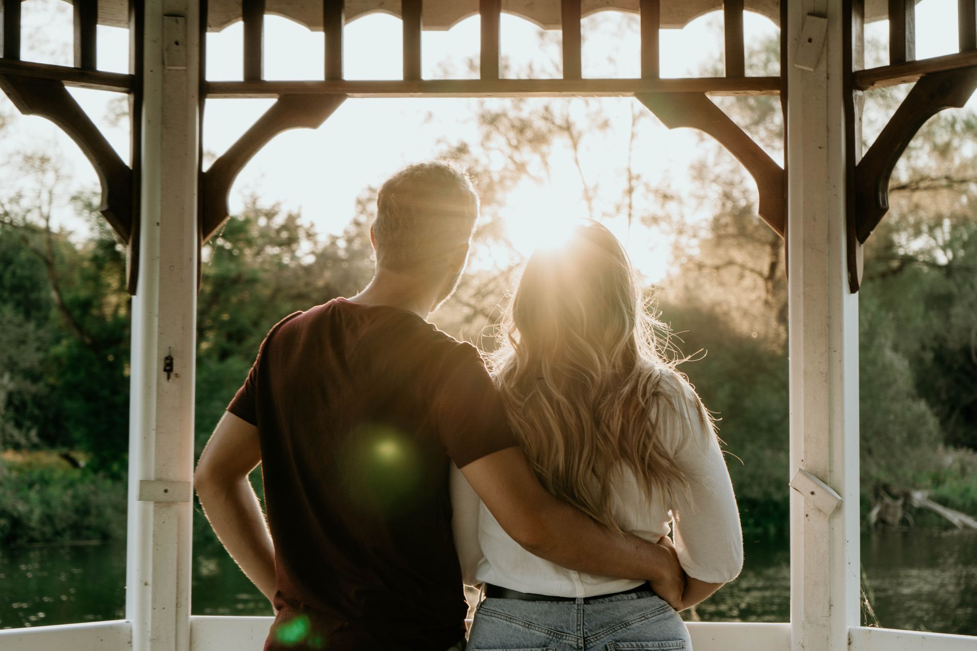 Couple embracing, watching sunset over a river from a white gazebo.