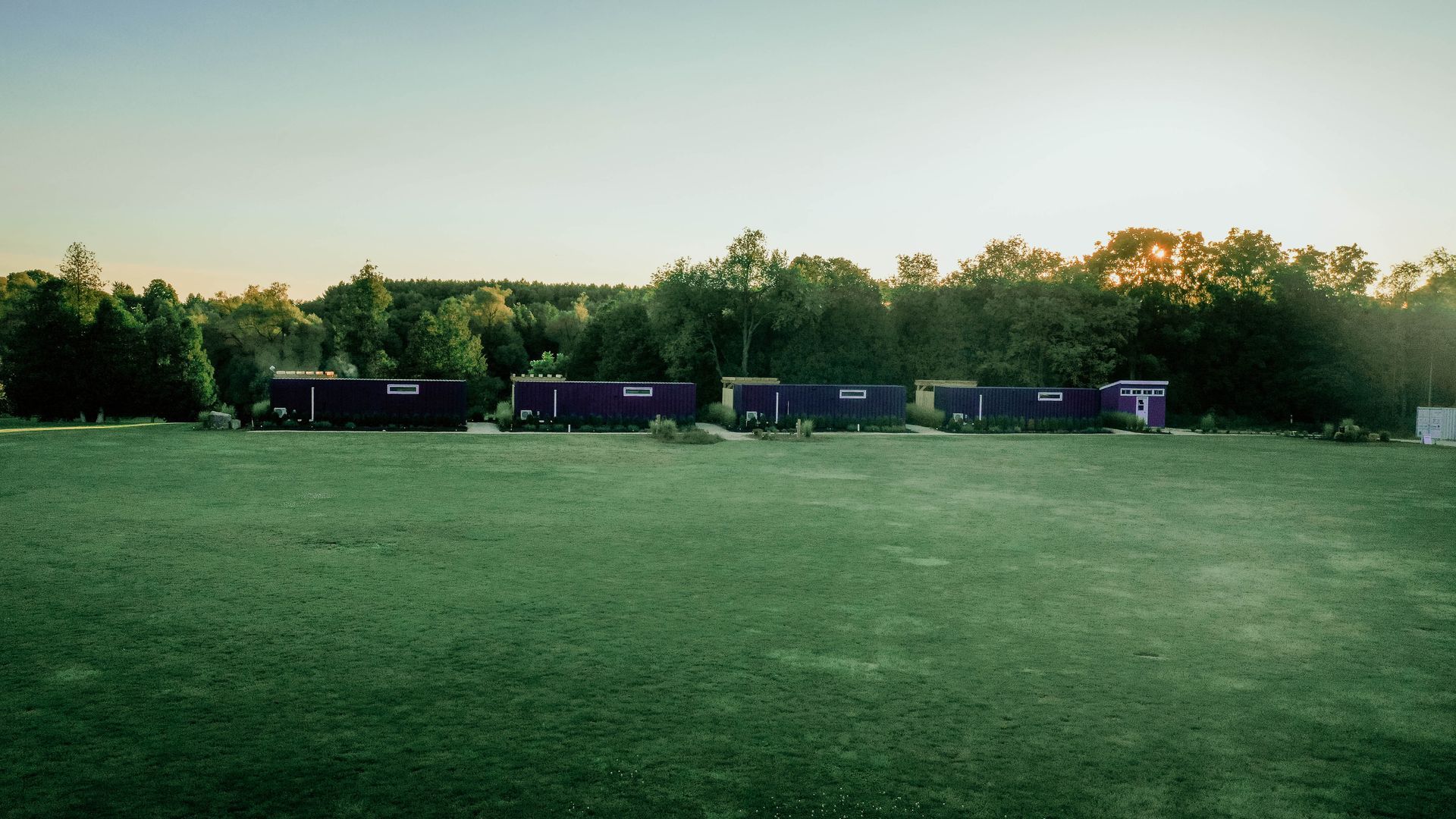 Row of dark cabins in a grassy field with trees in the background, lit by golden sunlight.