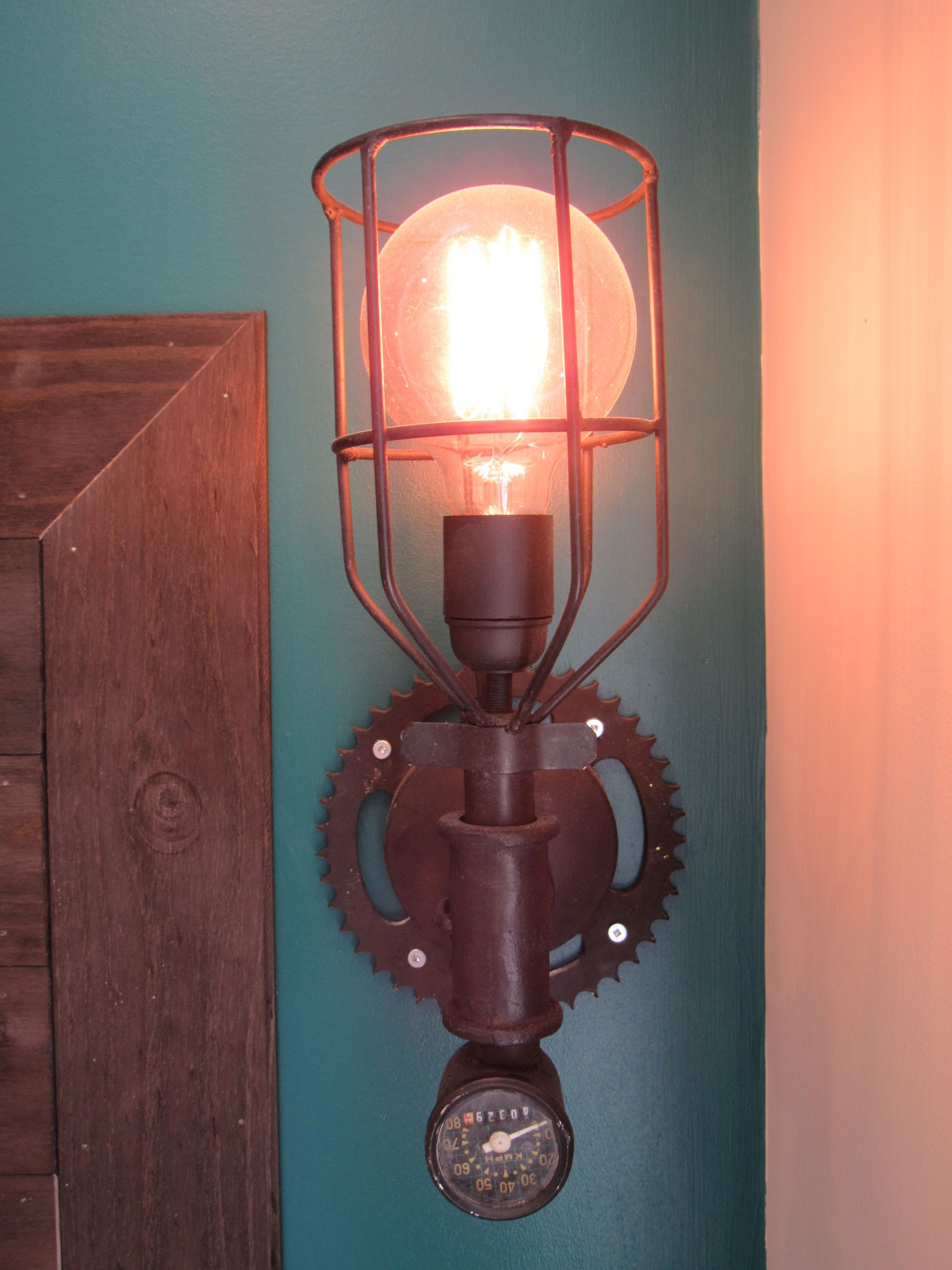 Steampunk-style wall sconce with a glowing Edison bulb, metal cage, and gear details mounted on a teal wall.