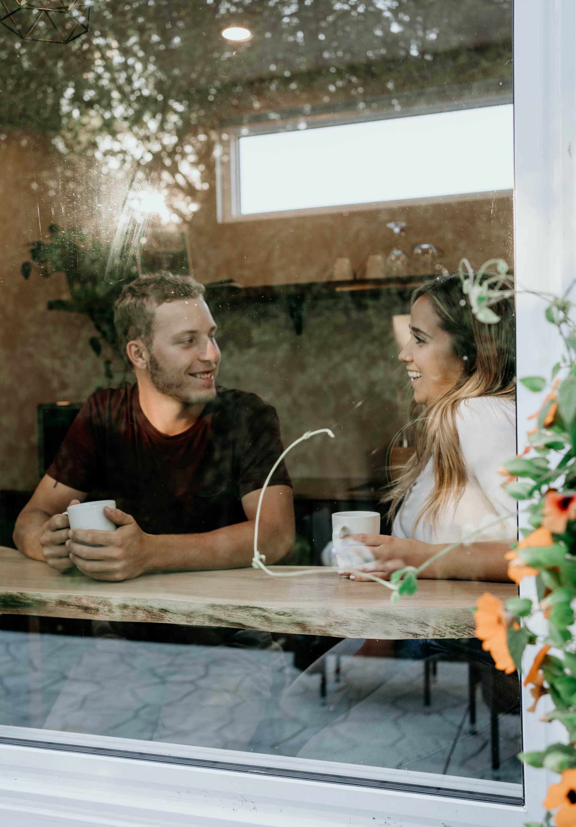 Couple seated at a cafe window, holding mugs, smiling, talking. Bright, natural light.