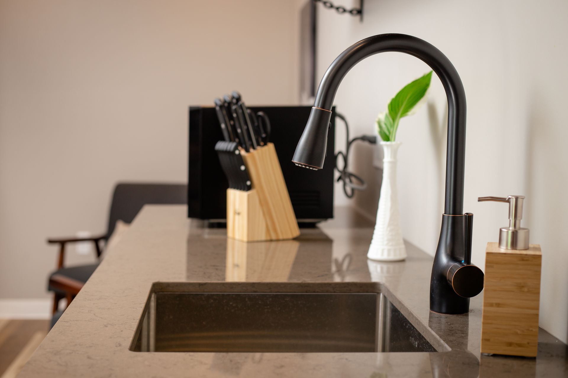 Black faucet, sink, counter, knives, dispenser, and white vase with plant on a kitchen counter.