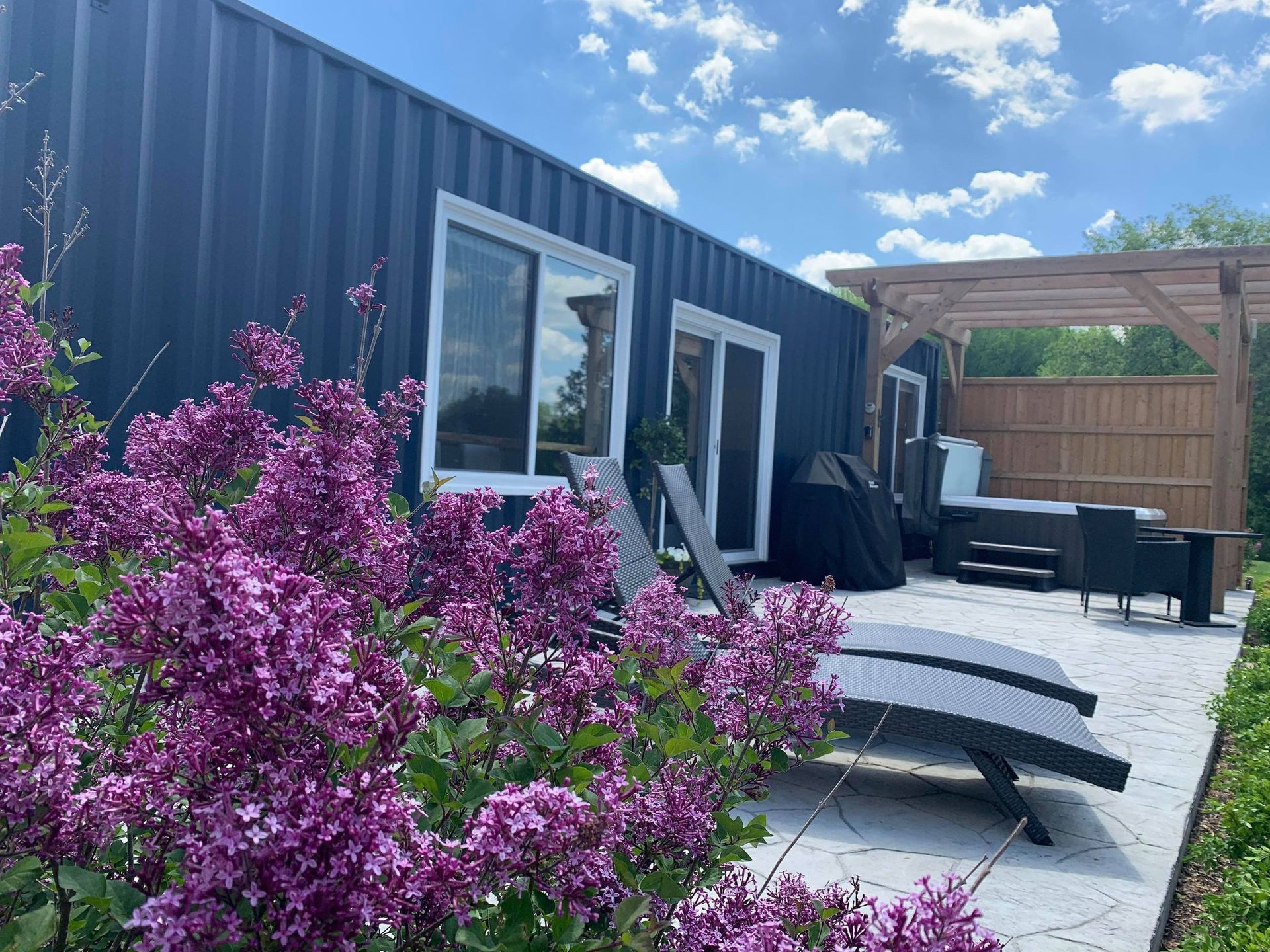 Dark blue shipping container home with patio, purple flowers, and lounge chairs on a sunny day.