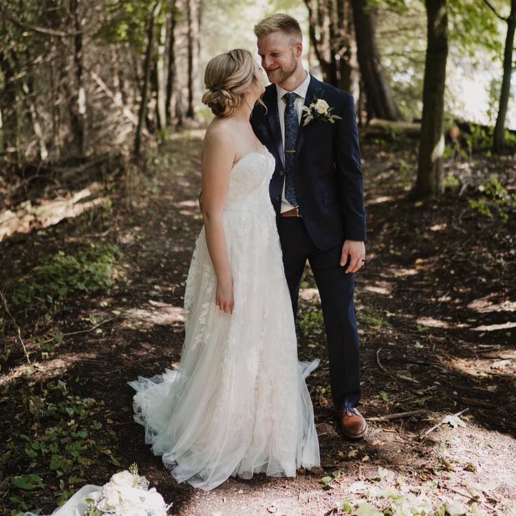 Bride and groom in formal attire smile at each other in a wooded area; bride wears white gown.