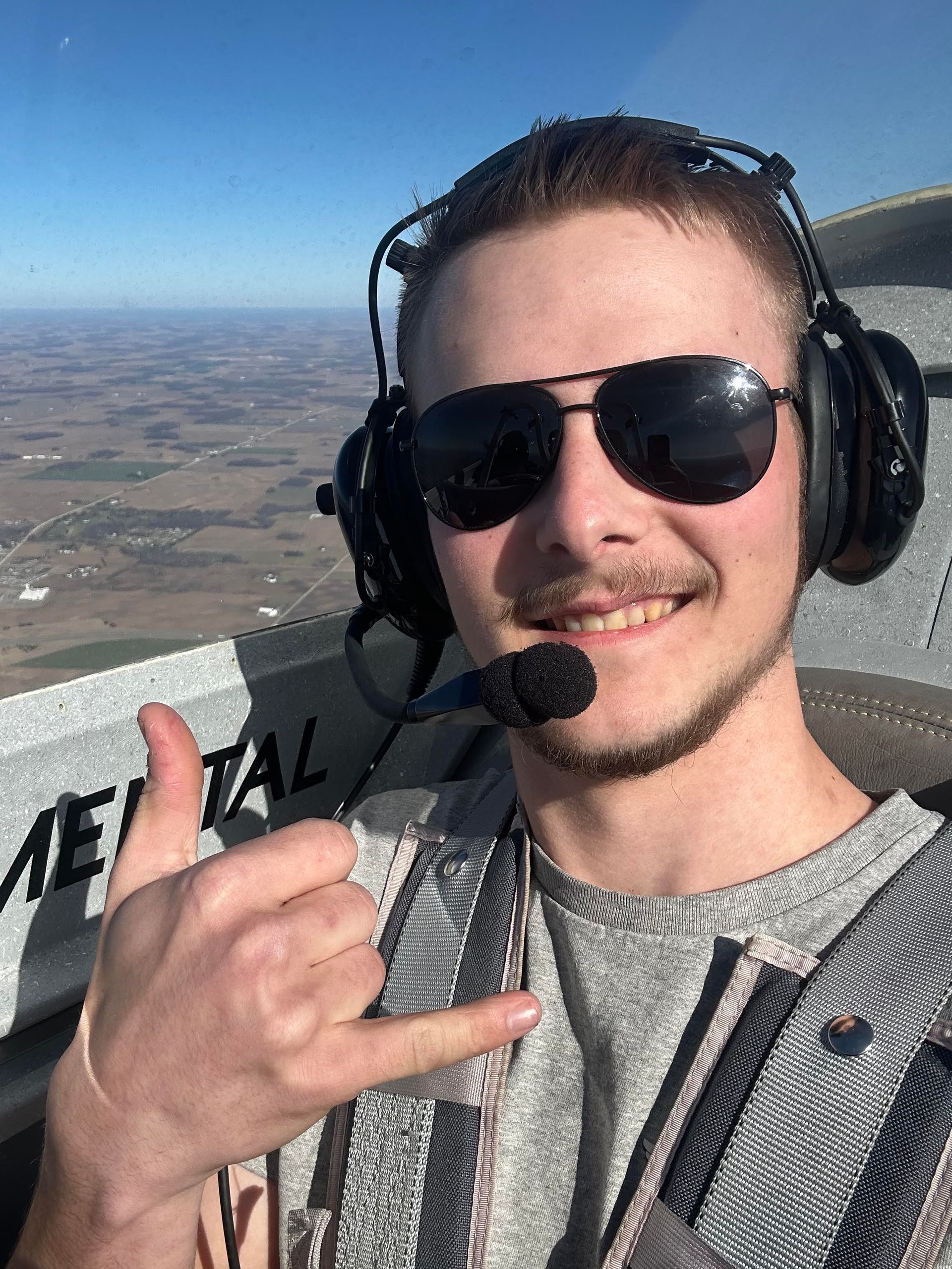 Person wearing sunglasses and headphones making a hand sign in an airplane cockpit over a landscape