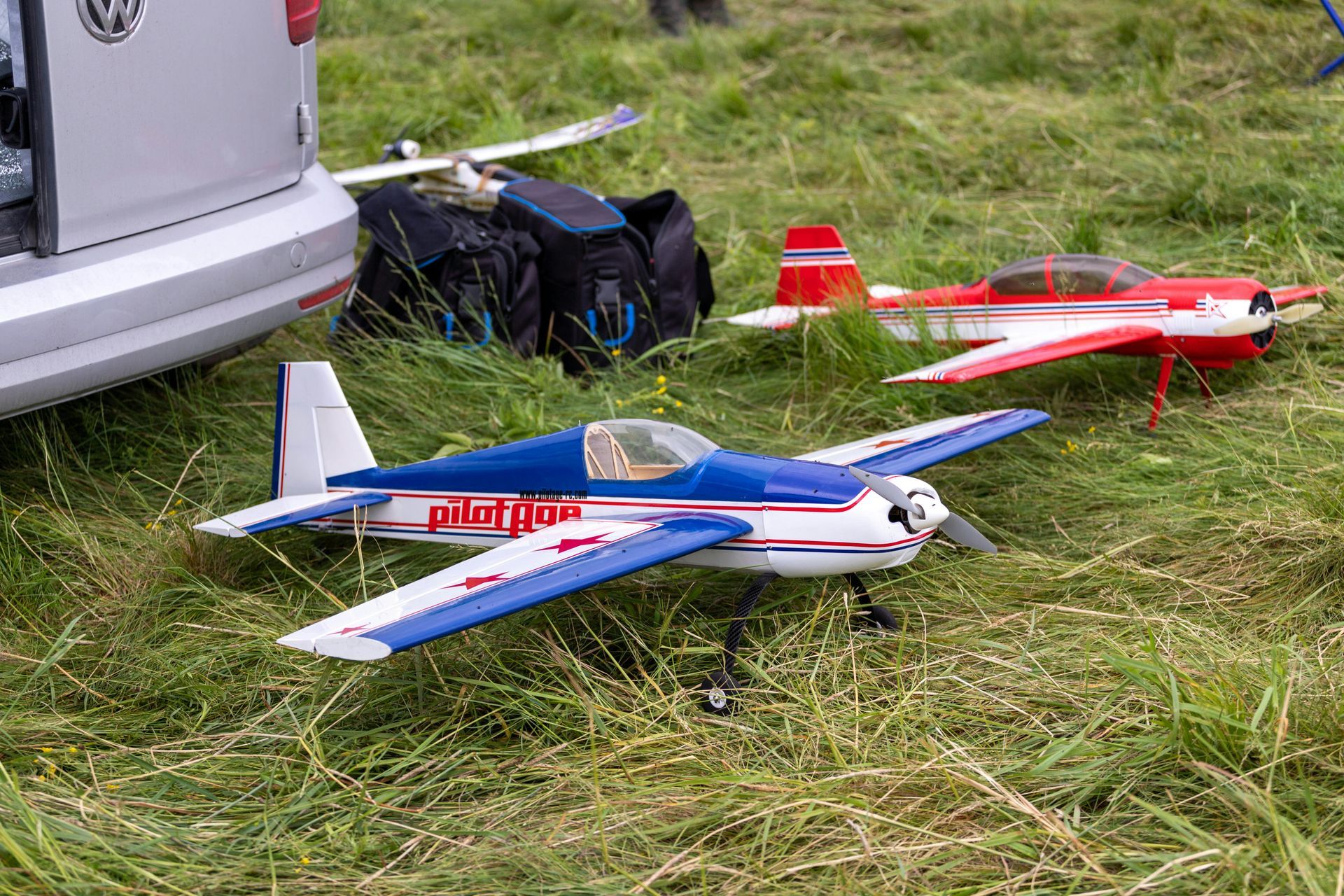 Two model airplanes on grass near a car. The closer one is blue and white; the other is red and white.