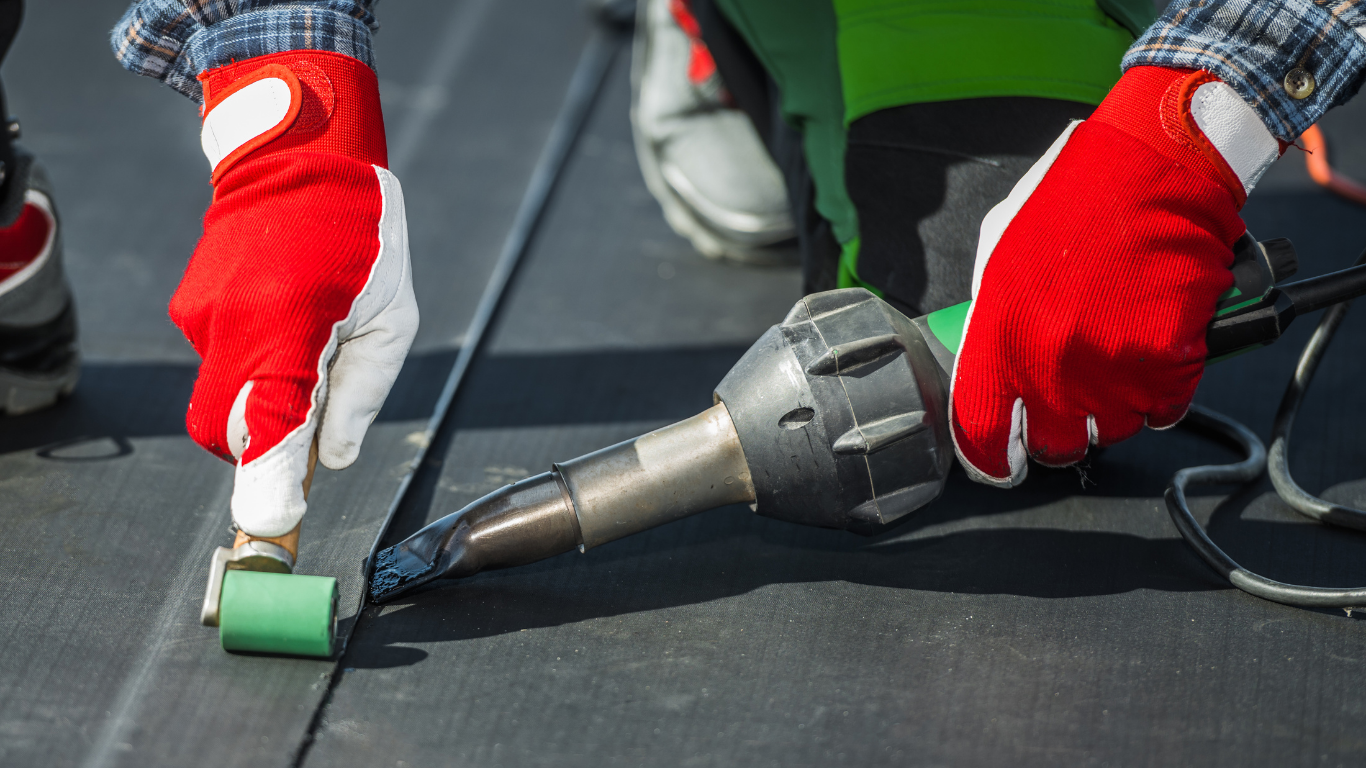 Hands in red gloves using a heat gun and roller to seal a black roofing seam.