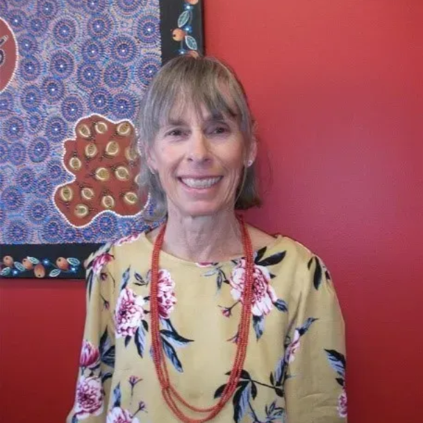 Woman with short gray hair, wearing a yellow floral blouse and red bead necklace, smiles in front of red wall with artwork.