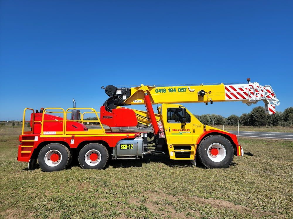 A Yellow And Red Truck With A Crane On The Back Is Parked In A Grassy Field — SureLift Crane Hire In Moranbah, QLD