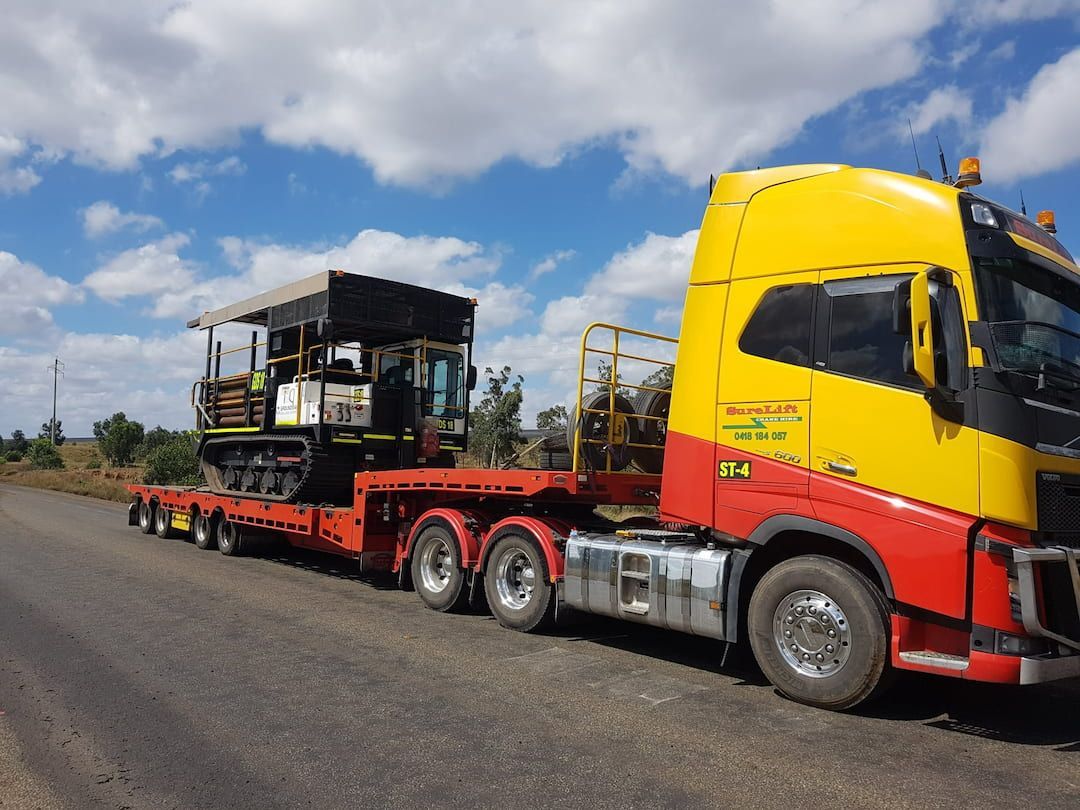 A Yellow And Red Semi Truck Is Carrying A Tractor Down A Road — SureLift Crane Hire In Mackay, QLD
